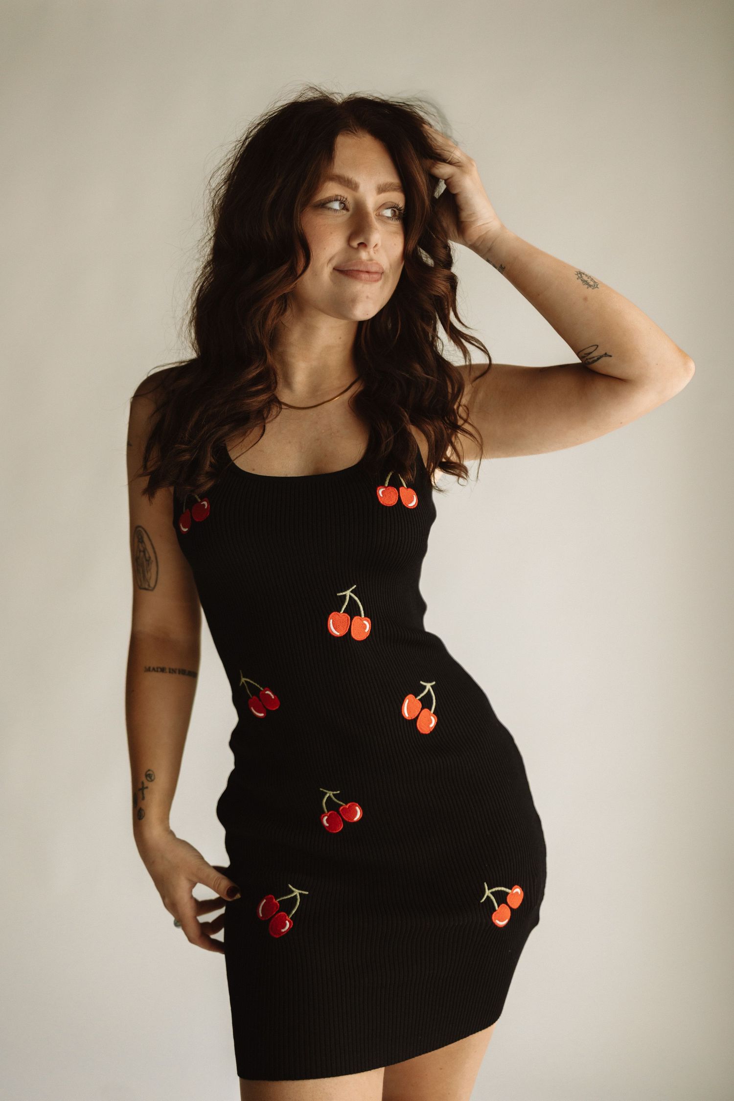 A black fitted dress adorned with red cherry appliques shown in a minimalist studio setting against a beige background.