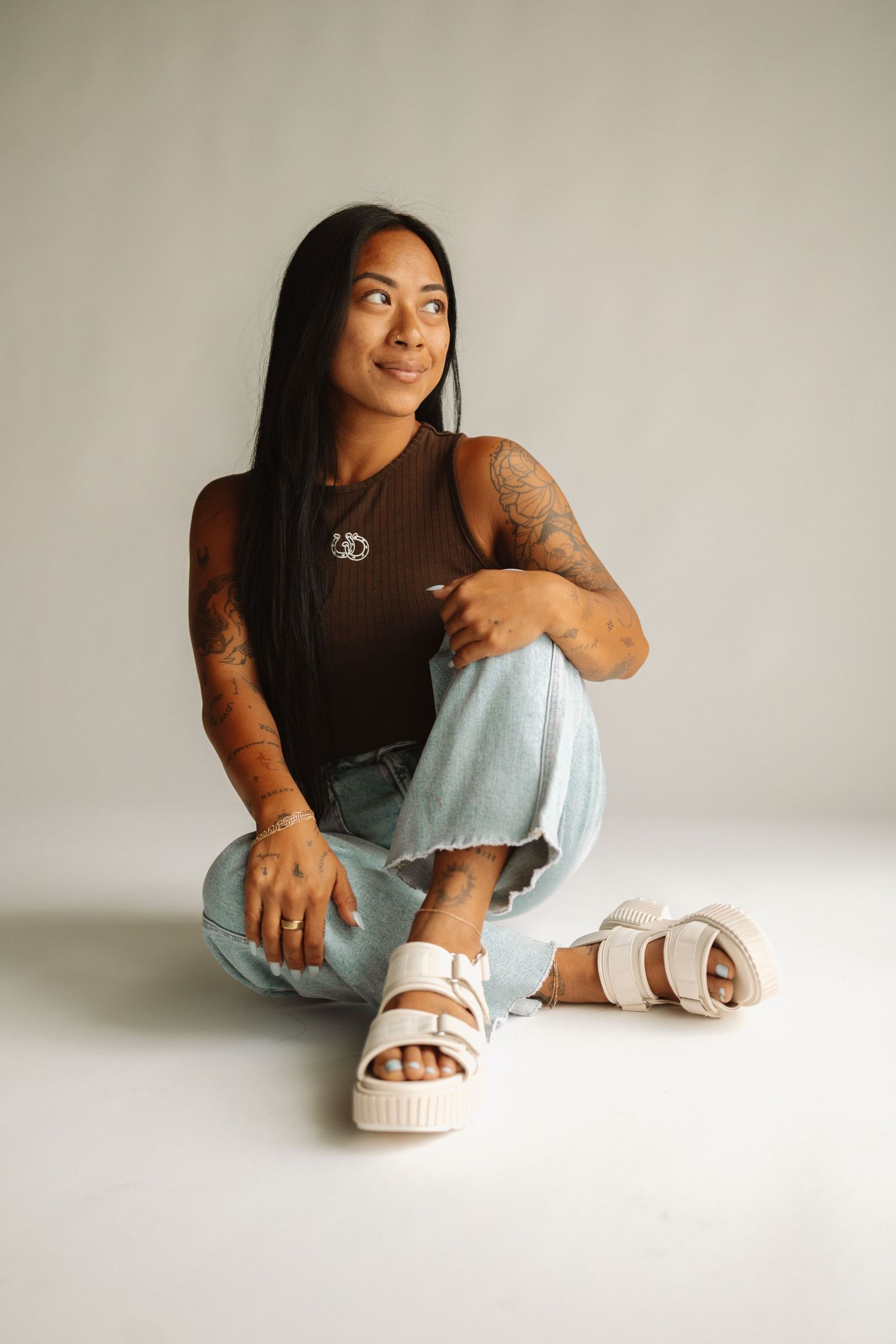 Person in athletic wear sits cross-legged on floor wearing casual white sandals and light denim jeans.