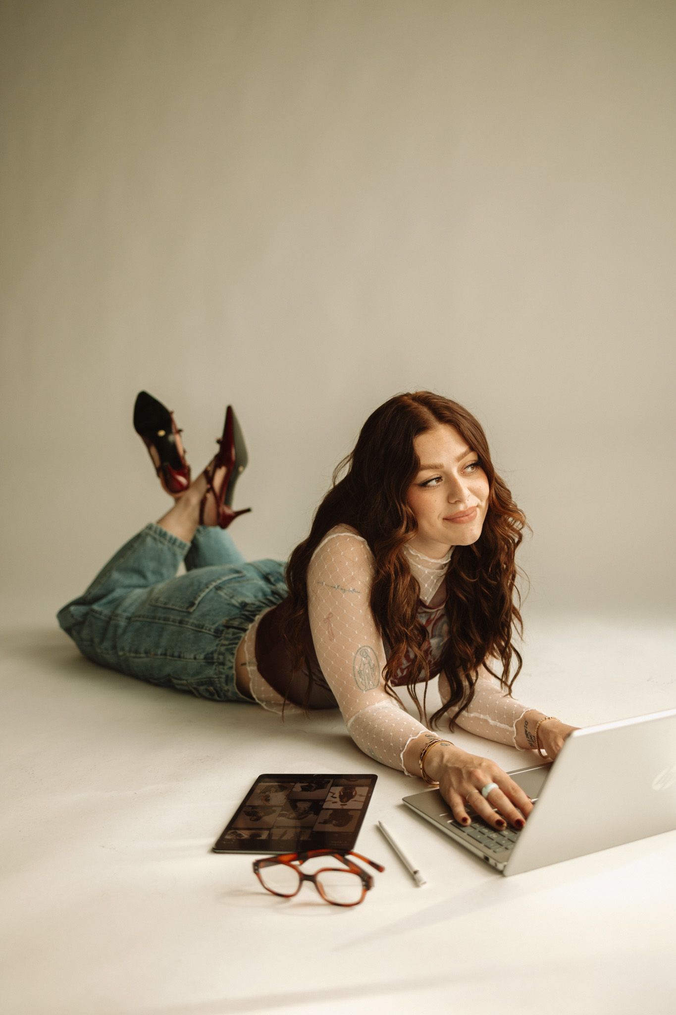 Woman lies casually on a white floor wearing jeans and a cream sweater while reading a magazine.