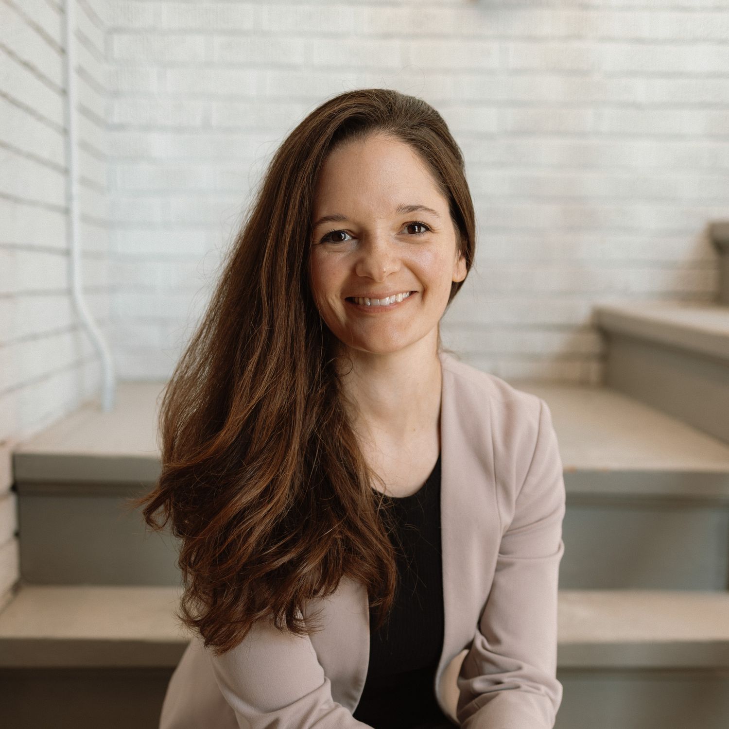 Professional in a beige blazer and black top sits on stairs with long brown hair and a warm smile.