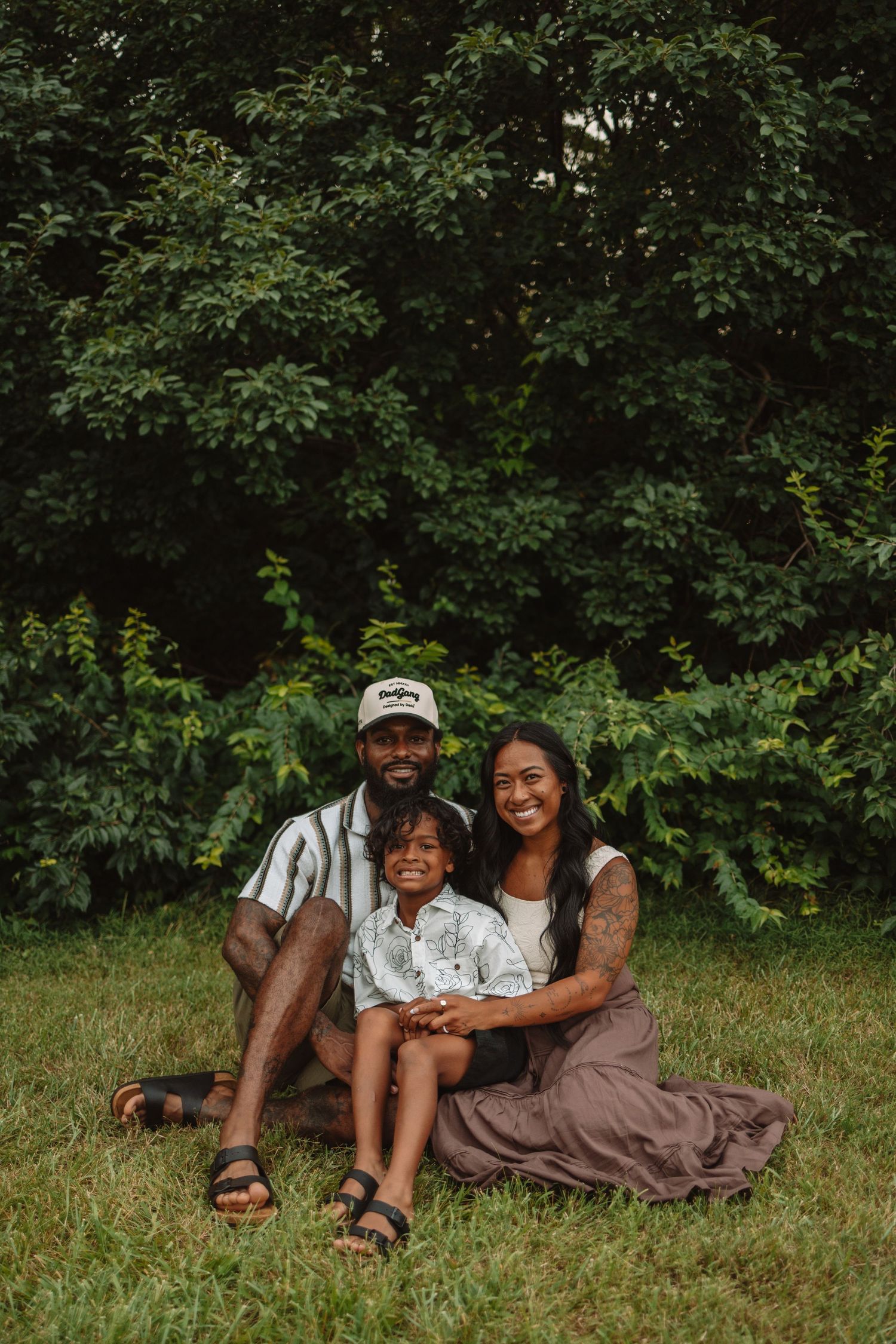 A family relaxes together on green grass during a warm summer day surrounded by lush foliage.