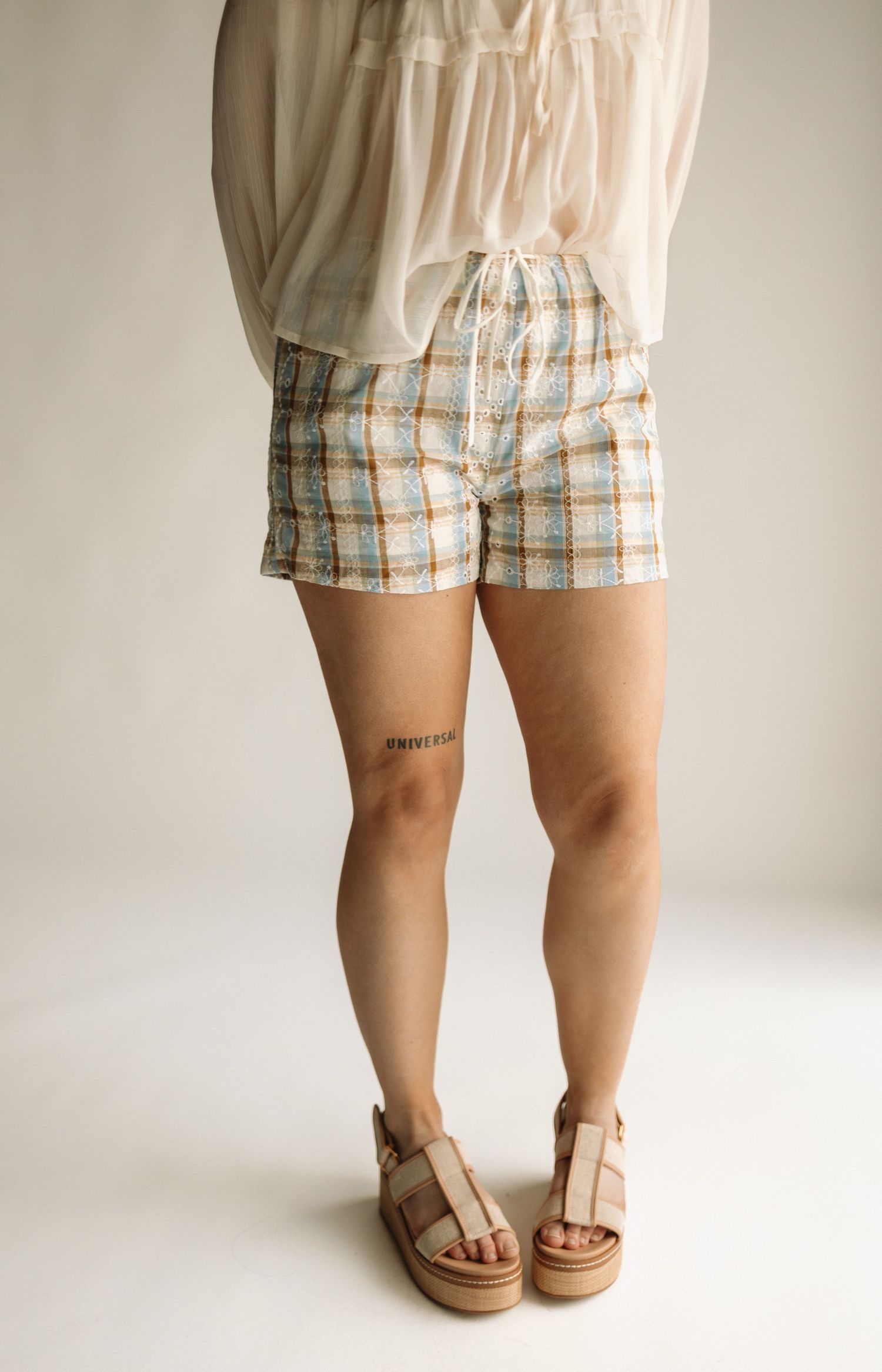 A person wearing tan sandals and plaid shorts stands on a beige floor against a light neutral background.