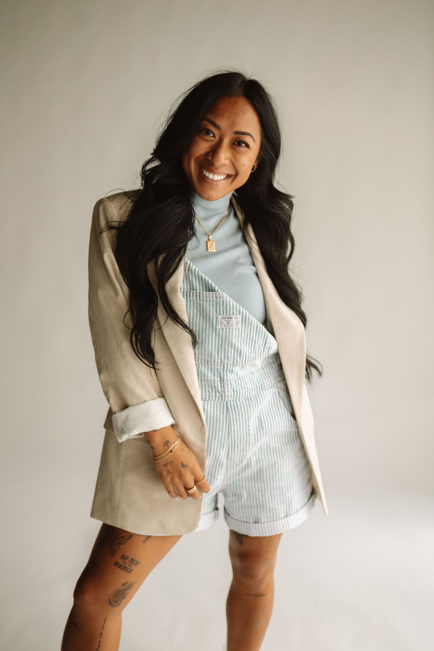 A person wearing a beige blazer and striped romper smiles warmly against a neutral background.