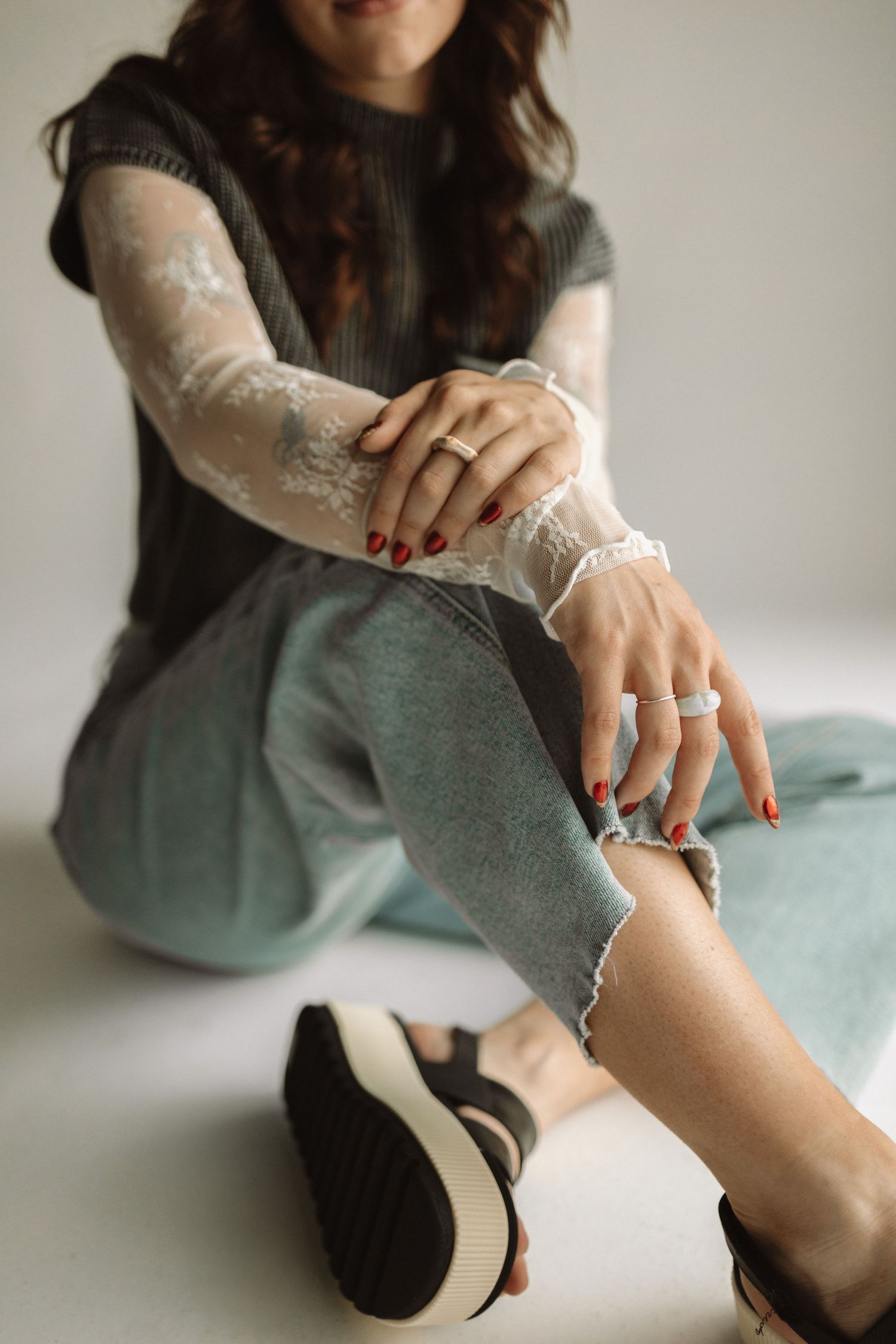 Hands with red nail polish and elegant white lace sleeves shown against sage green pants and black platform shoes.