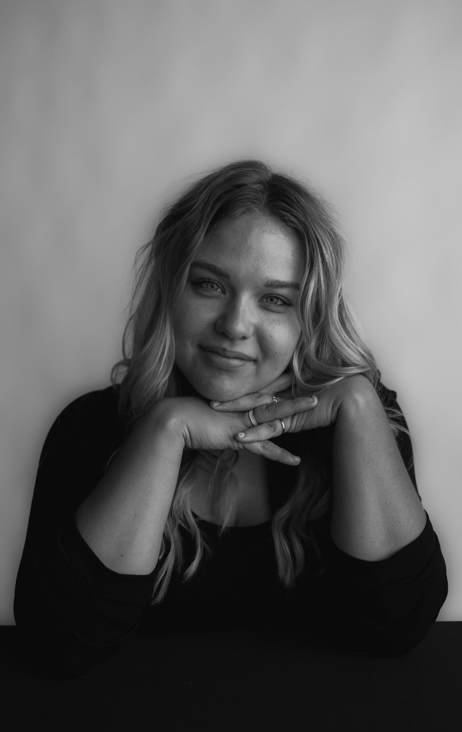 Black and white portrait photo showing a person resting their head on clasped hands while smiling warmly at the camera.