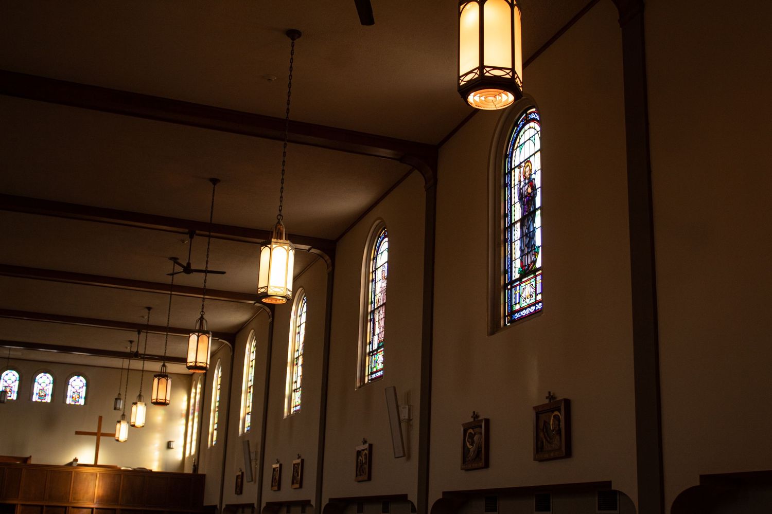 Illuminated pendant lights hang from exposed beams in a church interior with stained glass windows and gothic architecture.