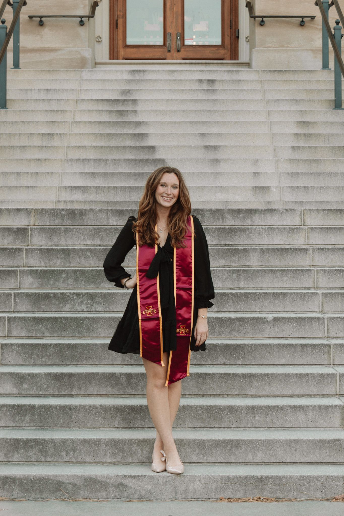 A graduate in a black dress and red graduation stole stands on concrete steps in front of a wooden door.