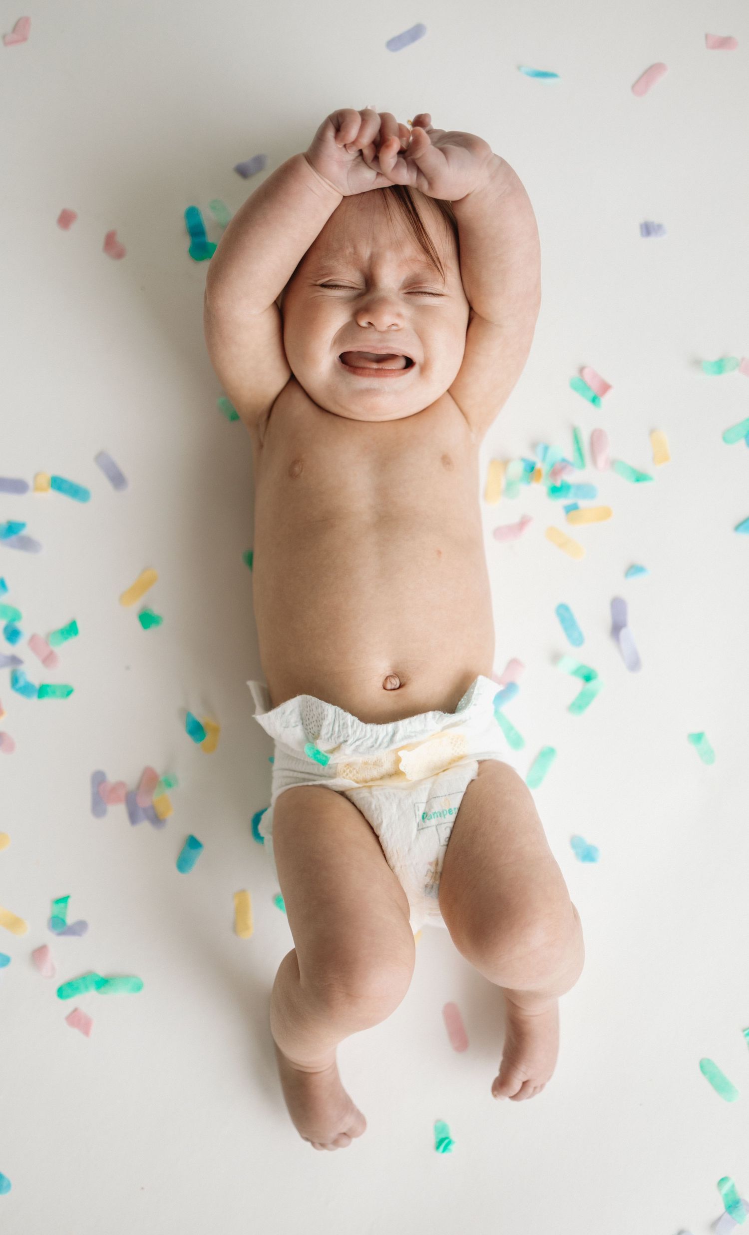 Baby lying on white background surrounded by colorful paper confetti in joyful celebration pose.