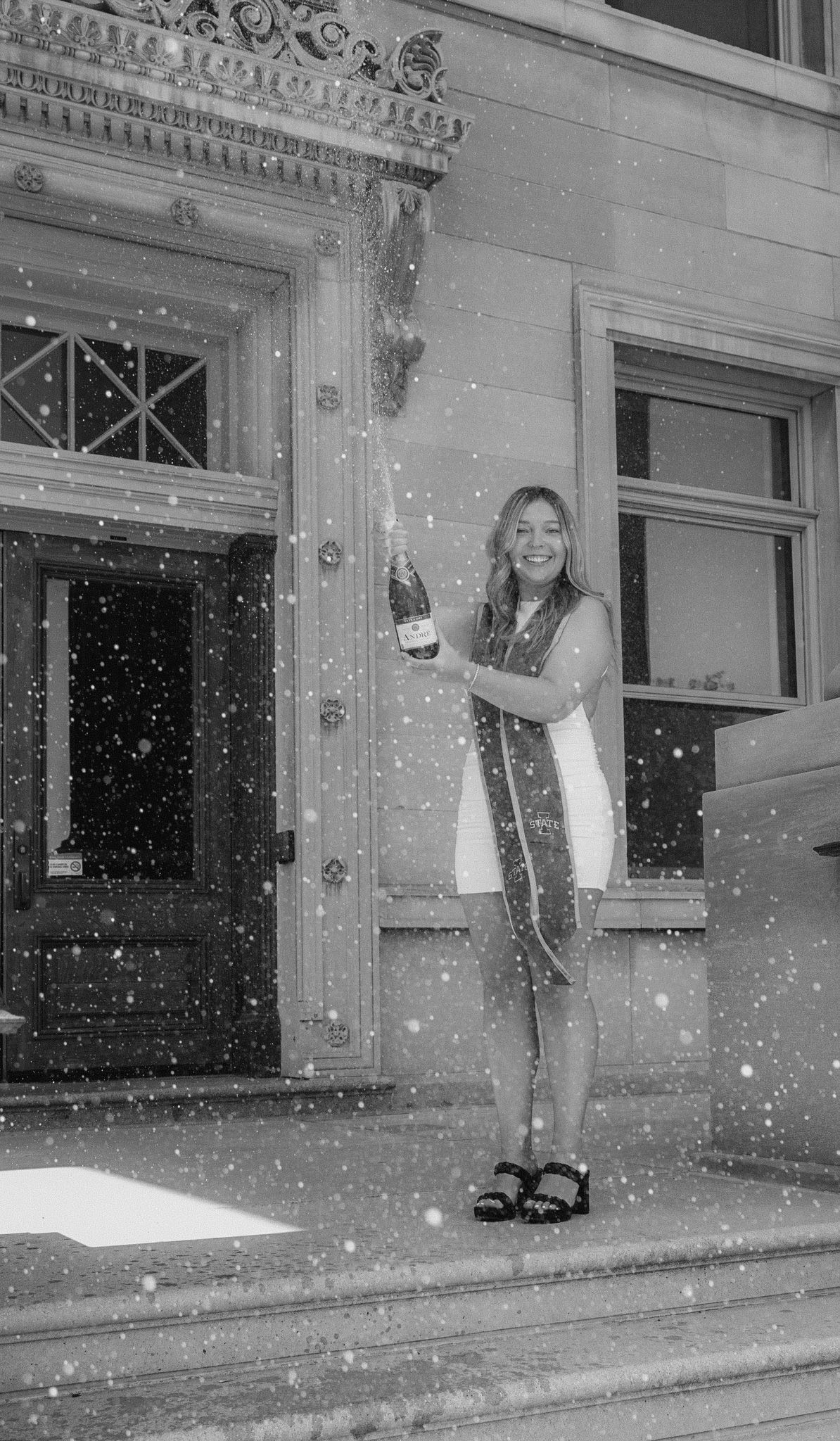 Graduate celebrates with champagne in front of a historic building entrance during snowfall.