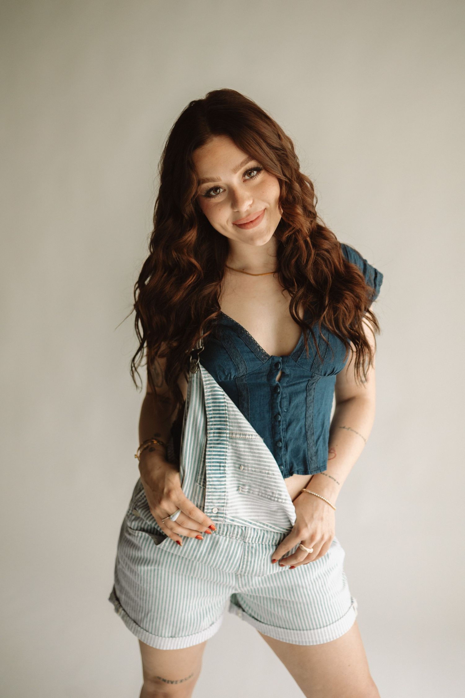 Young woman in denim romper overall shorts and navy top poses against beige wall in casual portrait setting.