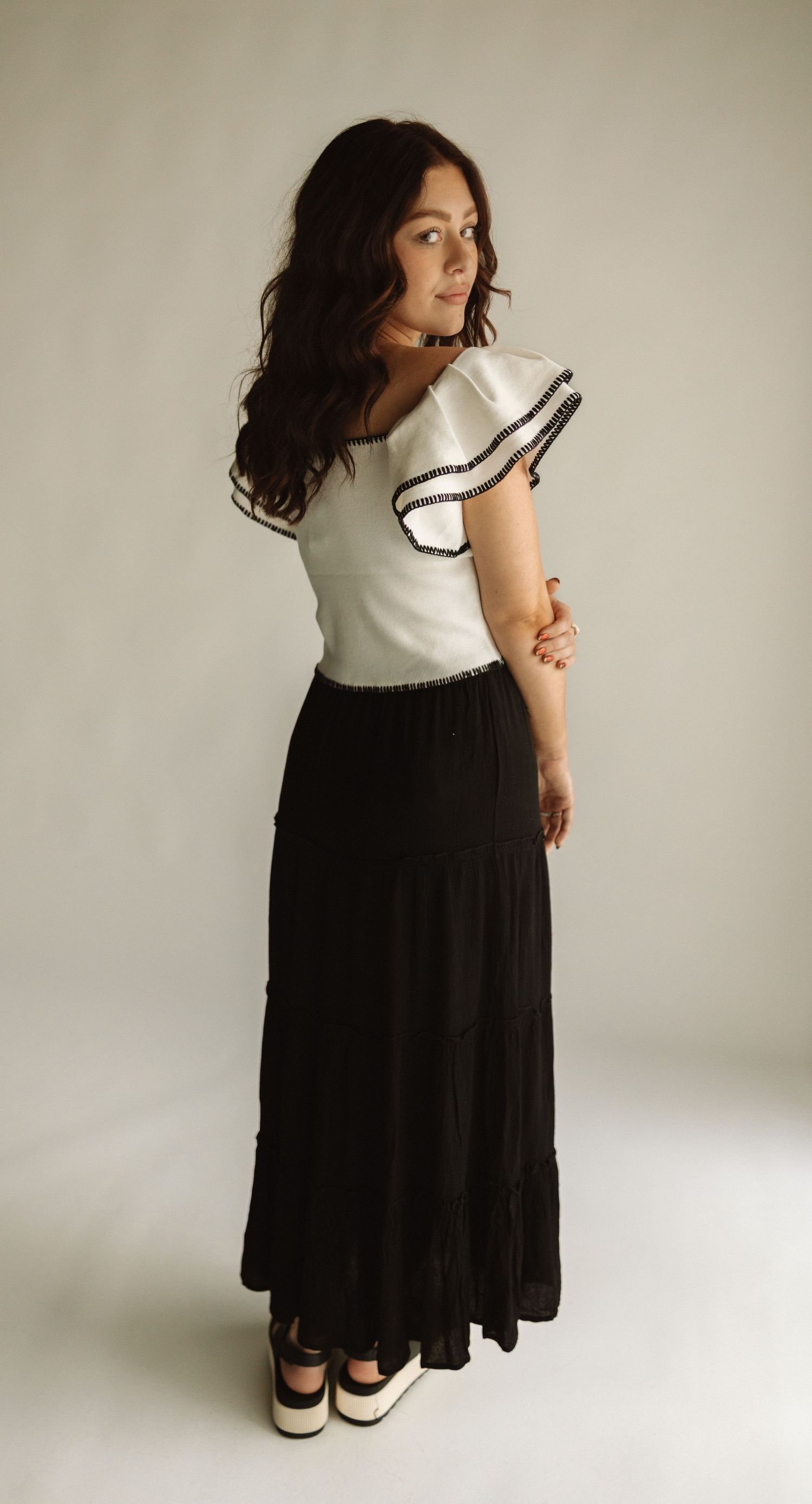 A person wearing a white ruffled top and long black maxi skirt poses against a neutral background.