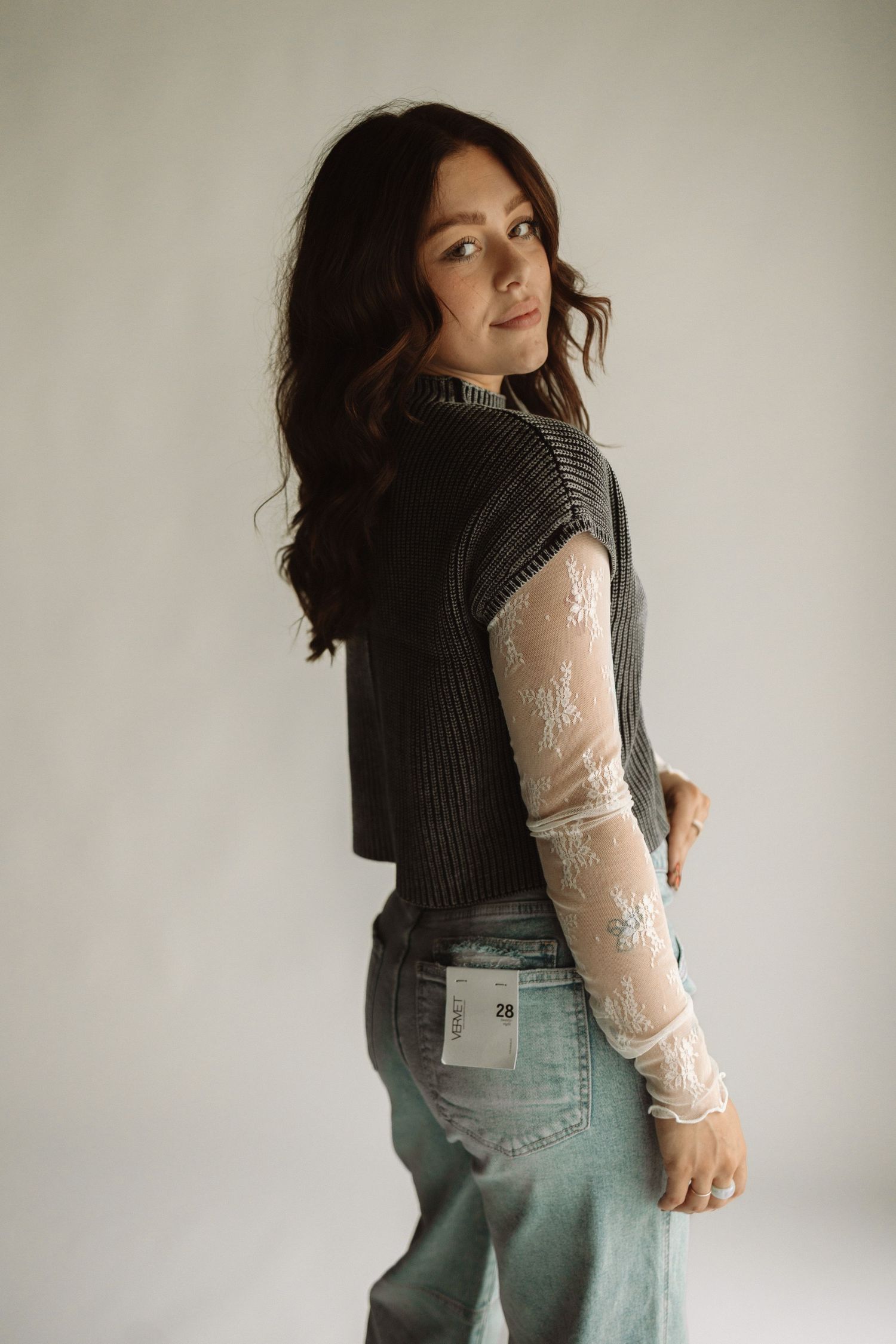 A woman poses in a dark knit vest layered over a sheer lace sleeve top and light denim jeans in a studio setting.