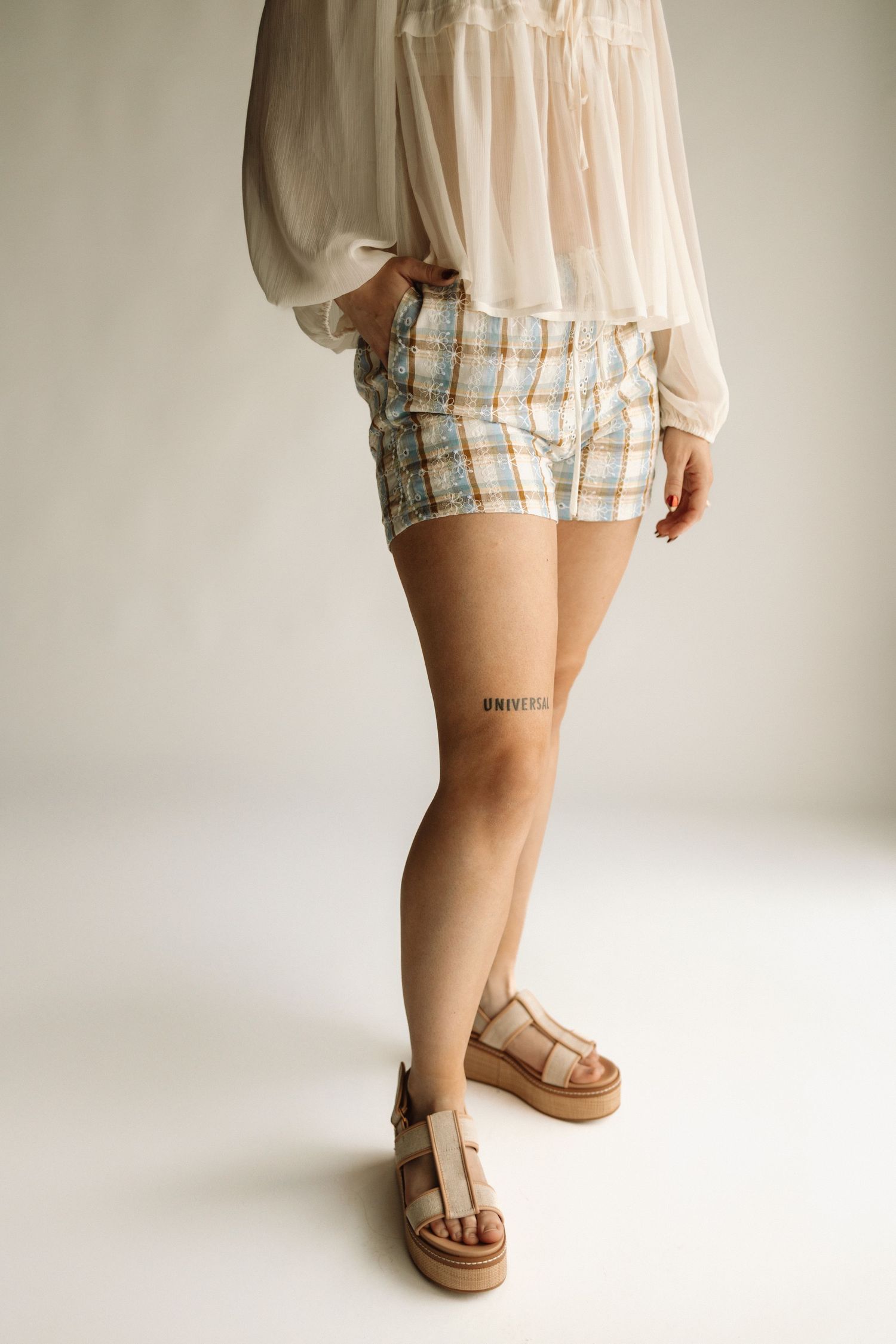 Side view of legs wearing plaid shorts, white blouse and beige sandals against white backdrop.