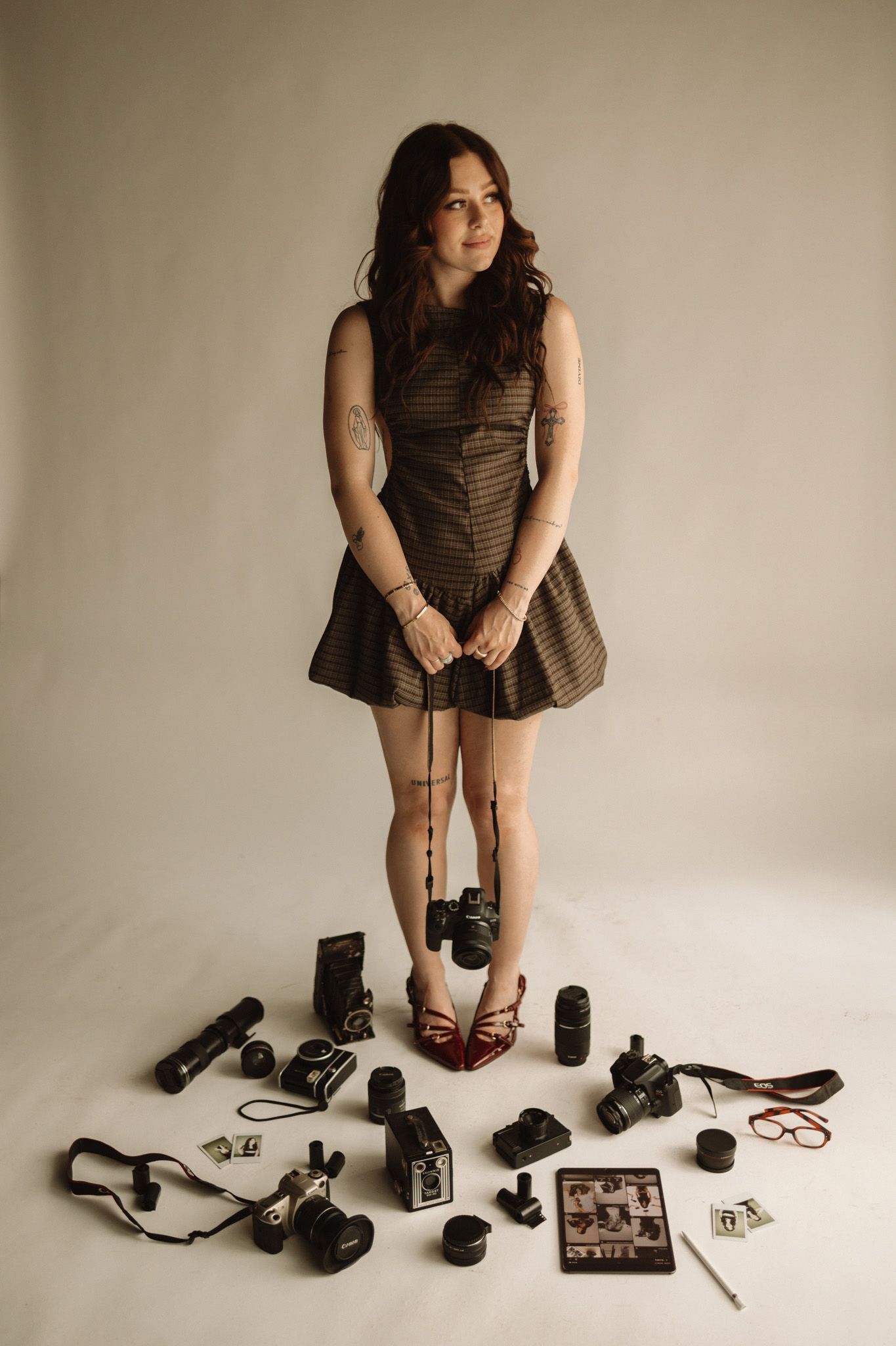 A photographer in a brown dress stands surrounded by camera equipment and lenses laid out on the floor.