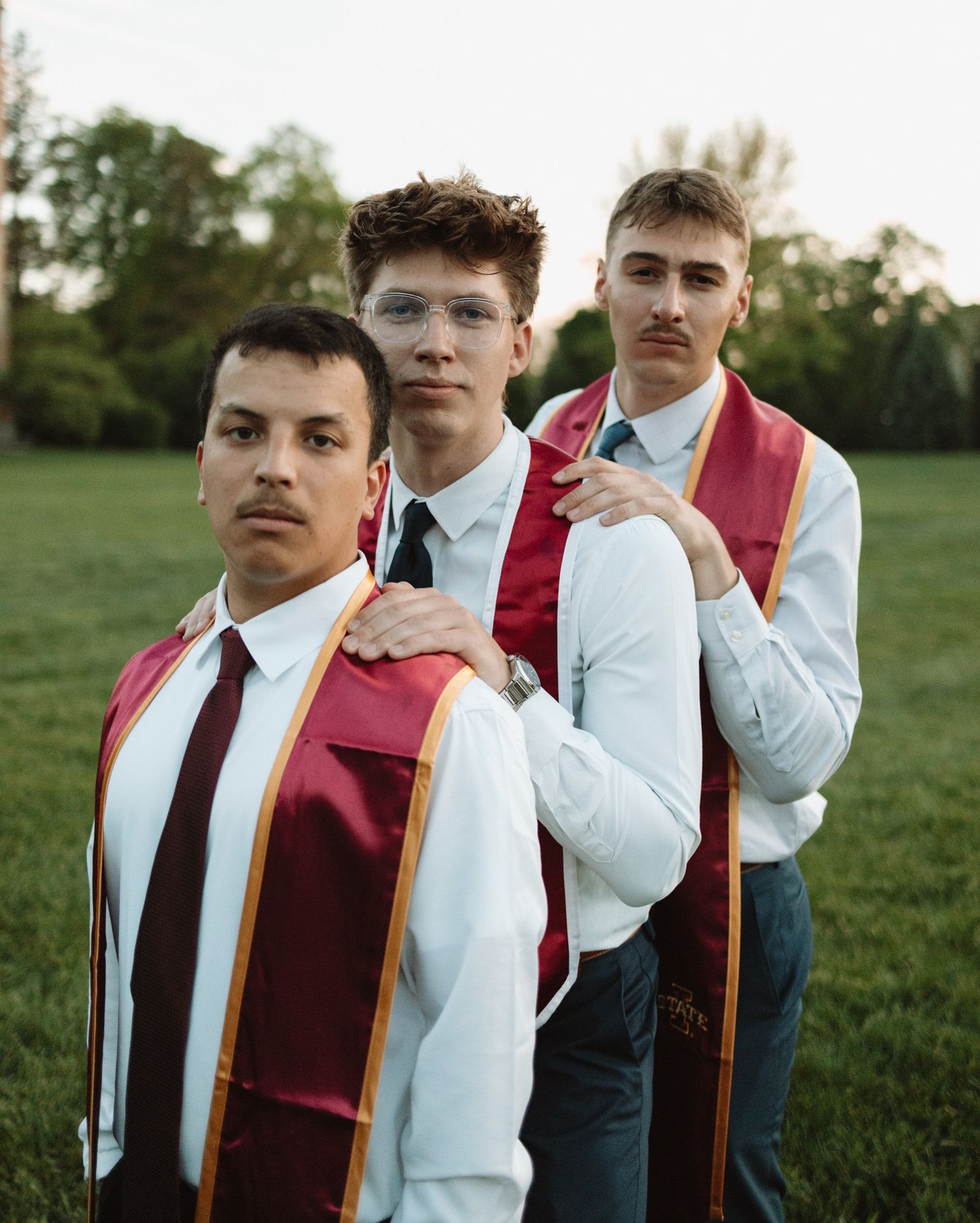 Row of graduates in formal attire and burgundy stoles pose together outdoors.