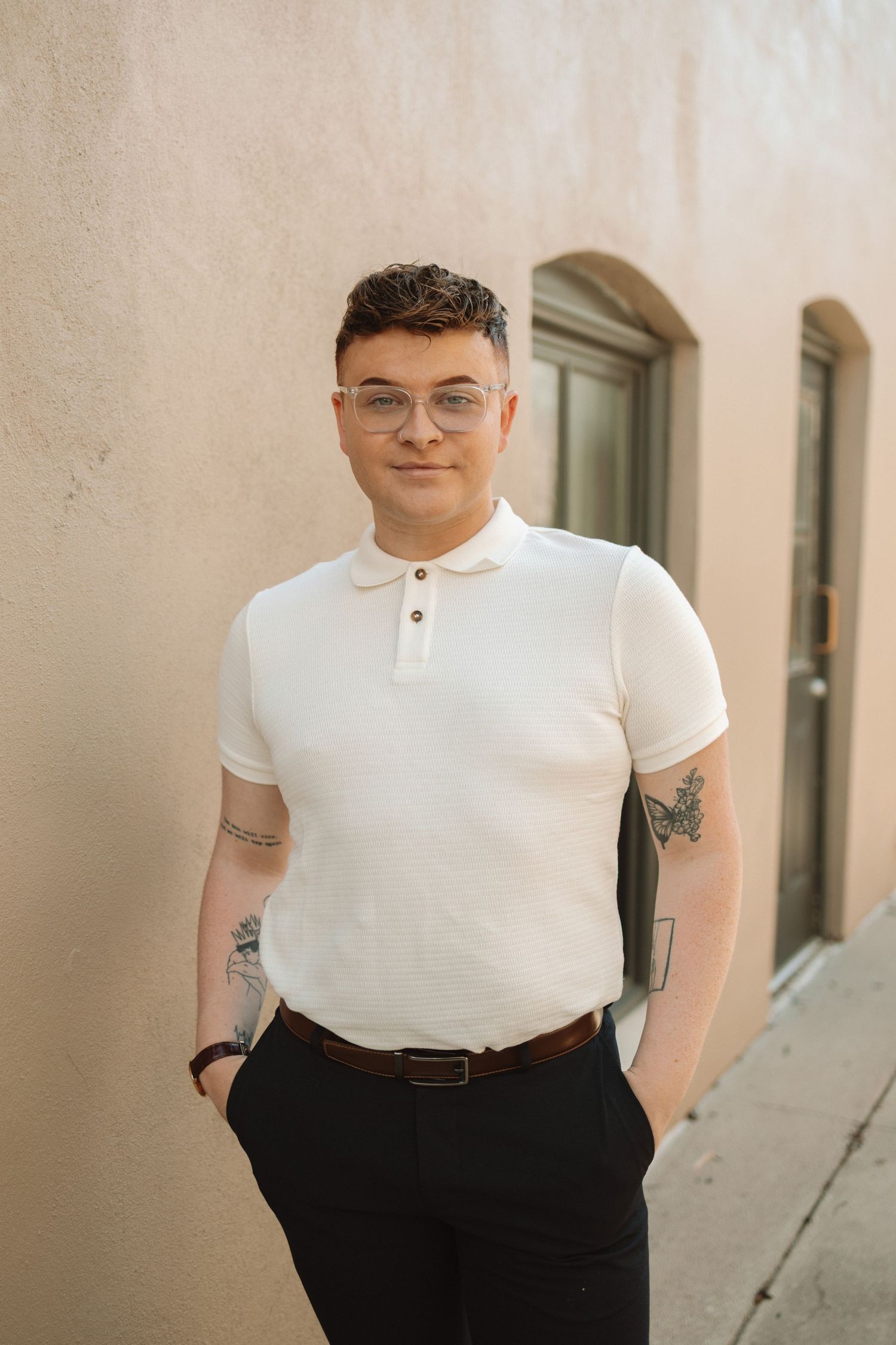 A person in a white polo shirt and dark pants stands against a beige wall with arched doorways.