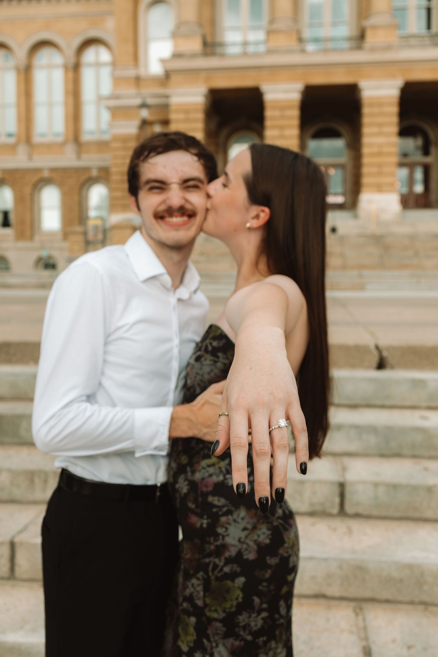 A couple shares a romantic moment on stone steps outside a historic building with tan colored walls.