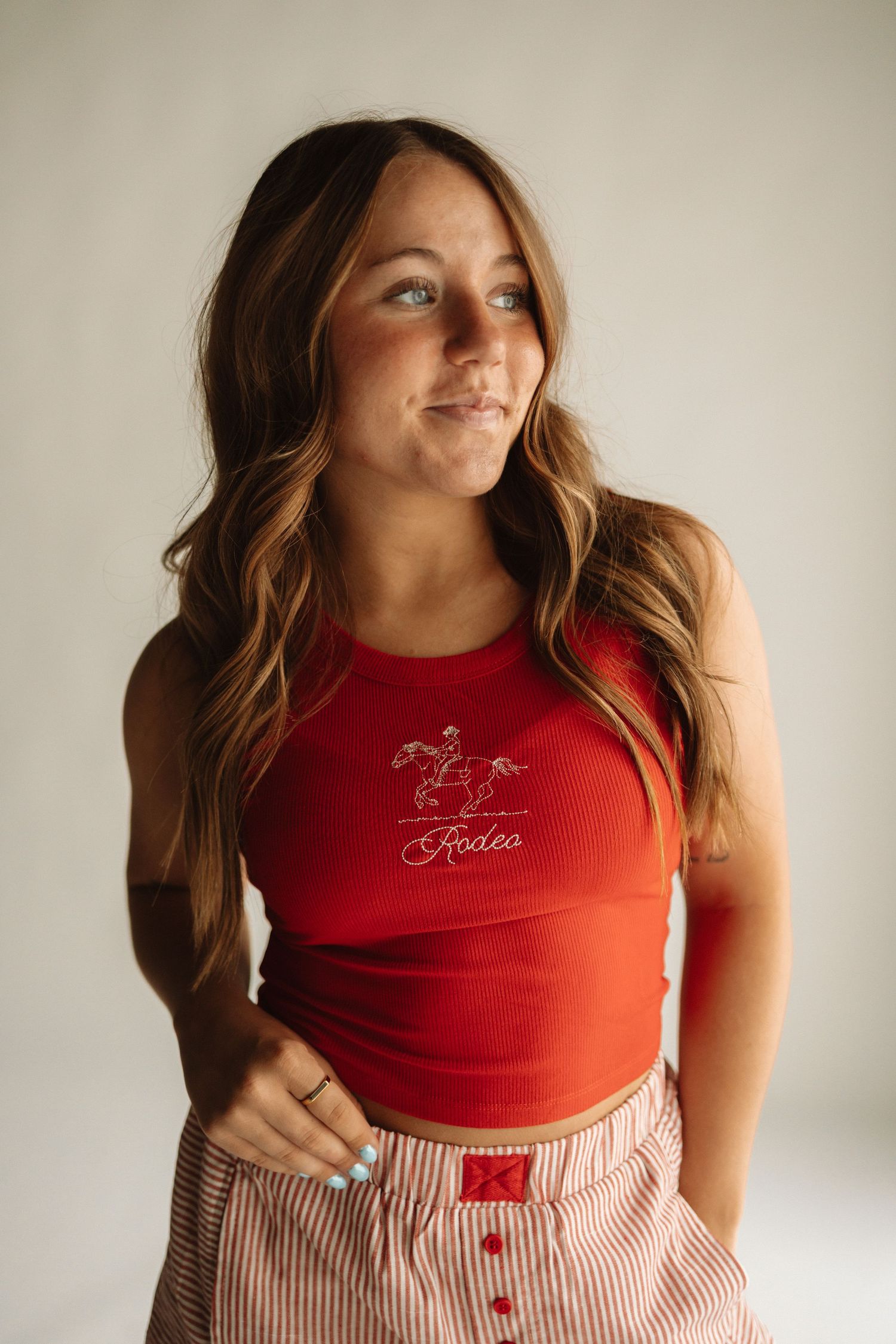 A woman in a red tank top and striped skirt poses for a portrait against a neutral wall.