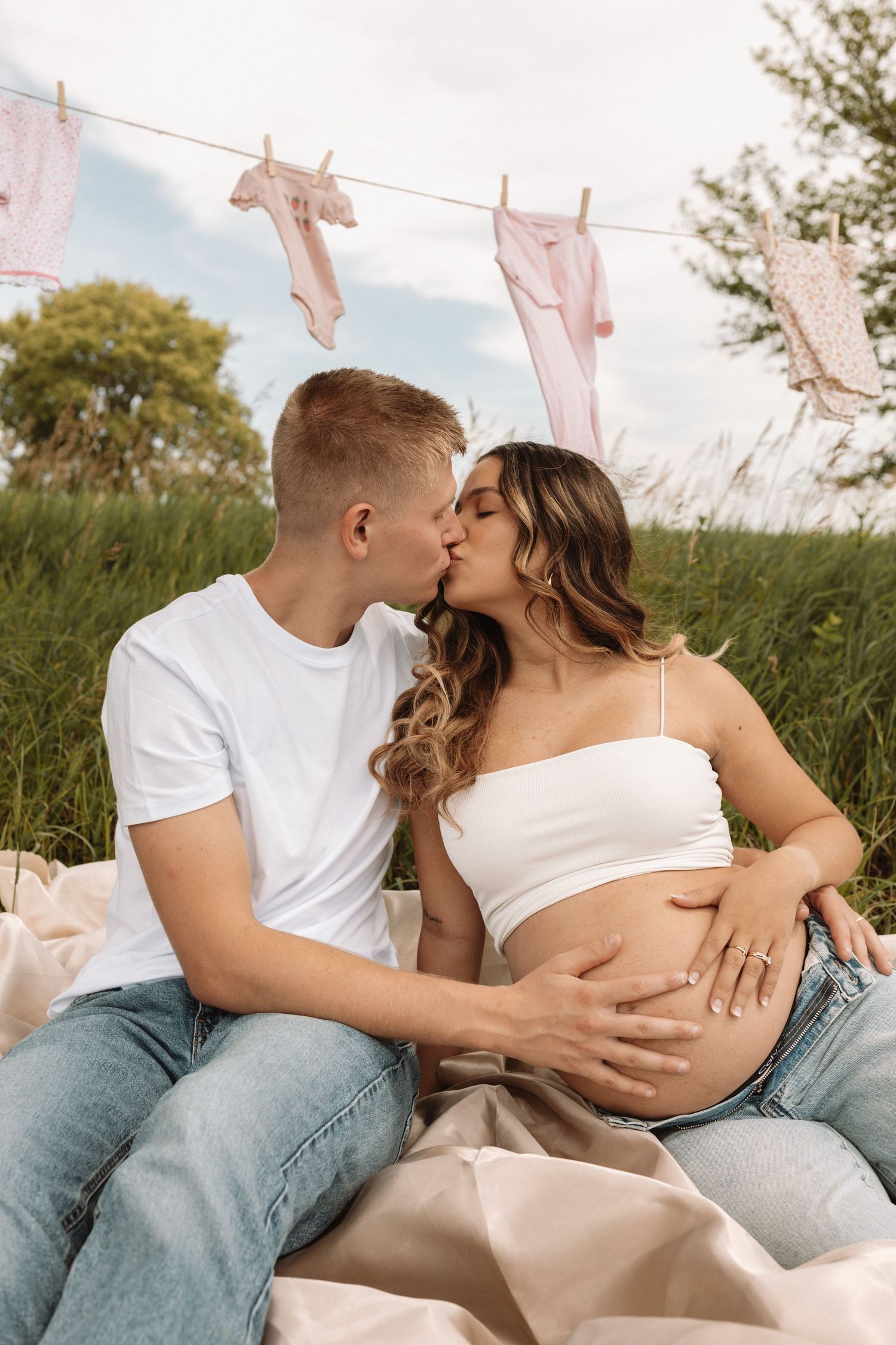 A pregnant couple sharing intimate moments on a blanket outdoors with baby clothes hanging on a clothesline above.