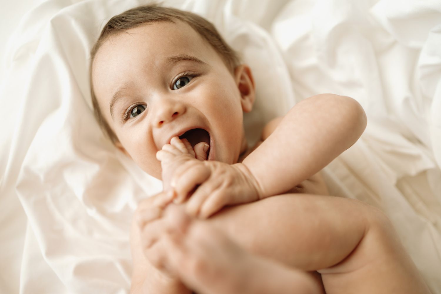A happy baby lies on soft white fabric while laughing and holding their feet.
