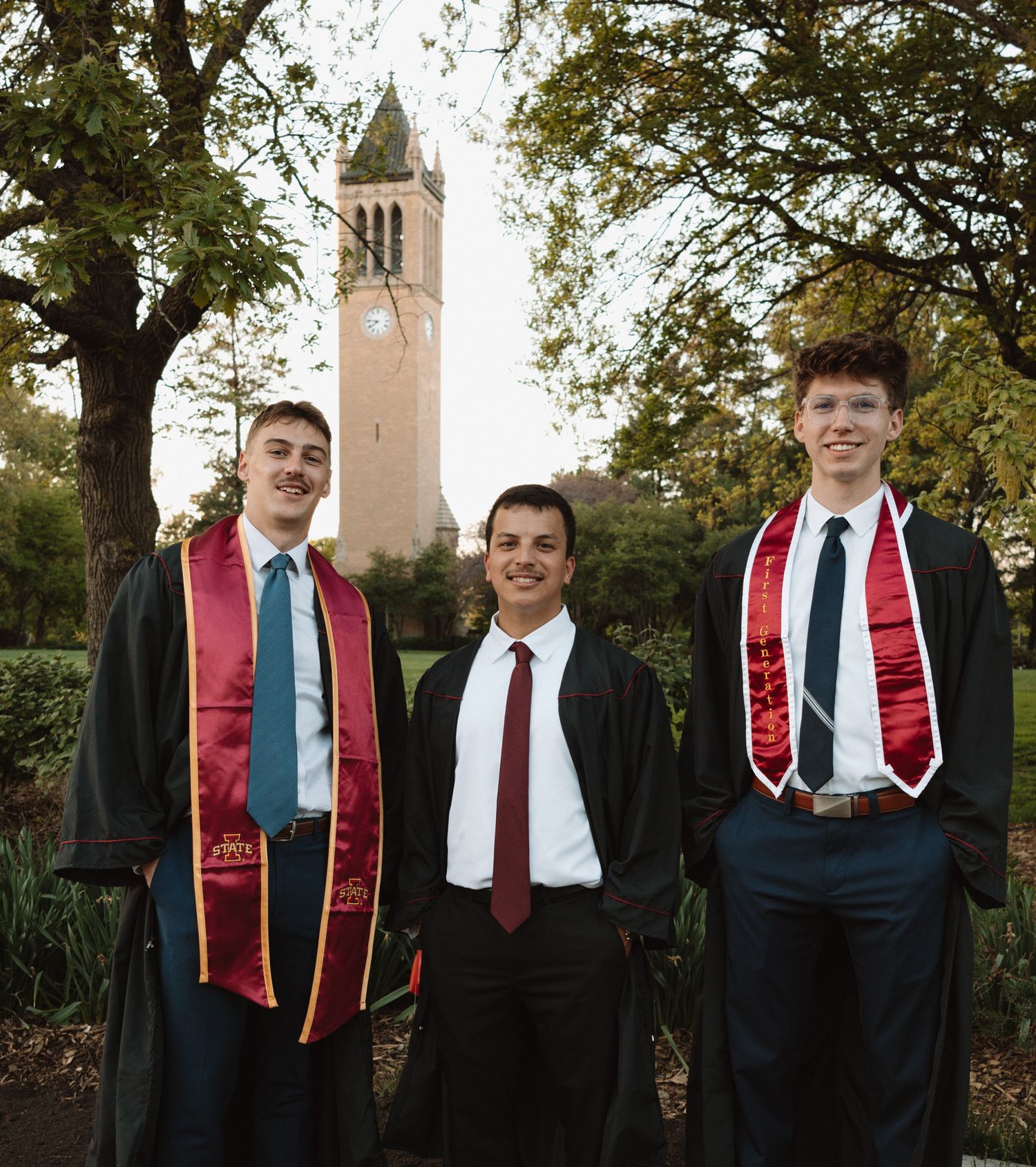 Three graduates pose together in front of a campus bell tower on graduation day.