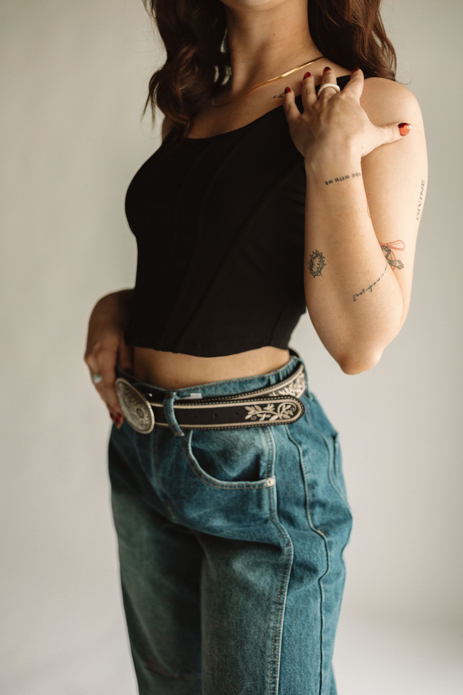 Person wearing black crop top and blue jeans with western belt showing small minimalist arm tattoos.