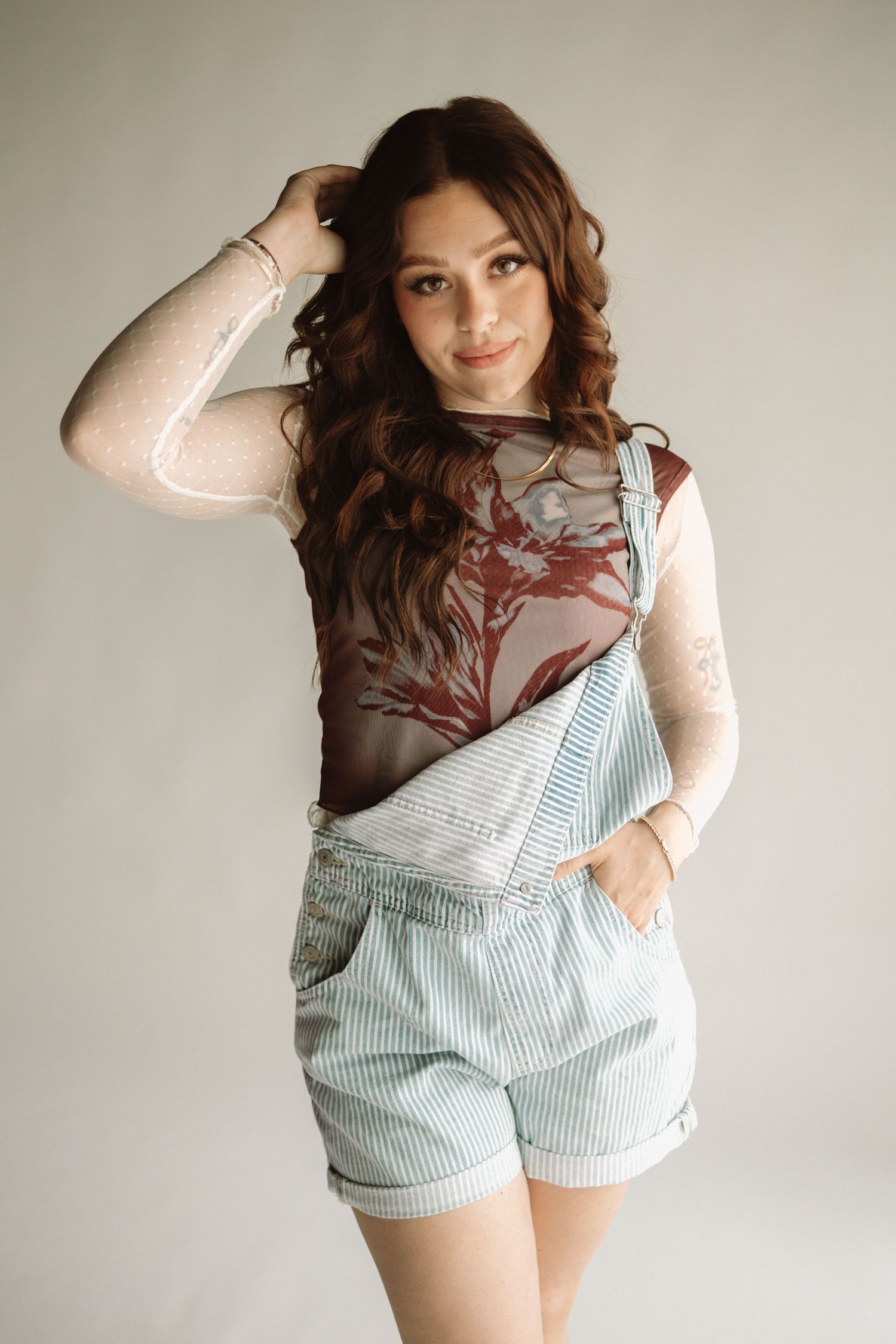 A person poses in striped light blue overalls shorts and a sheer patterned long-sleeve top against a neutral wall.