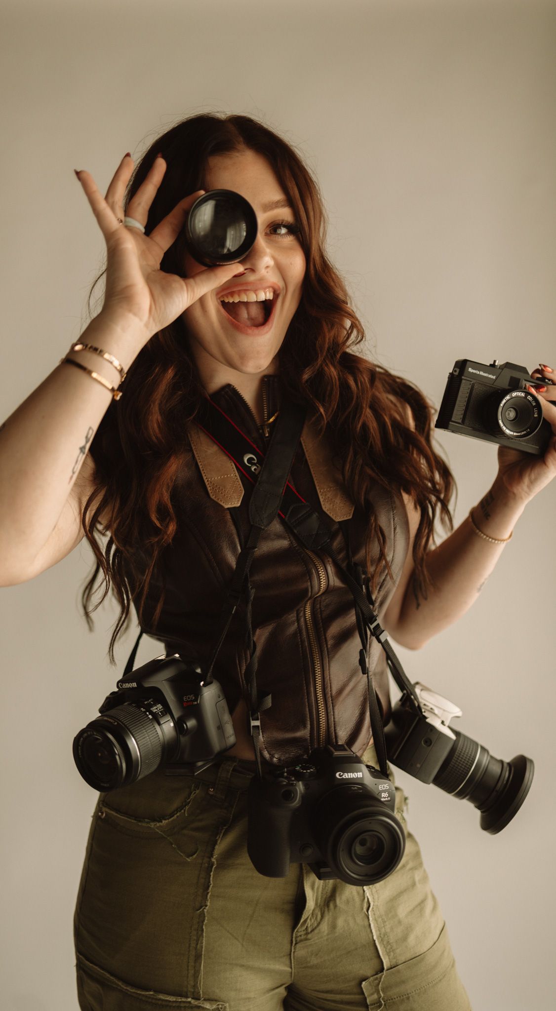 Photographer wearing dark top and olive pants poses playfully with multiple cameras in a studio setting.
