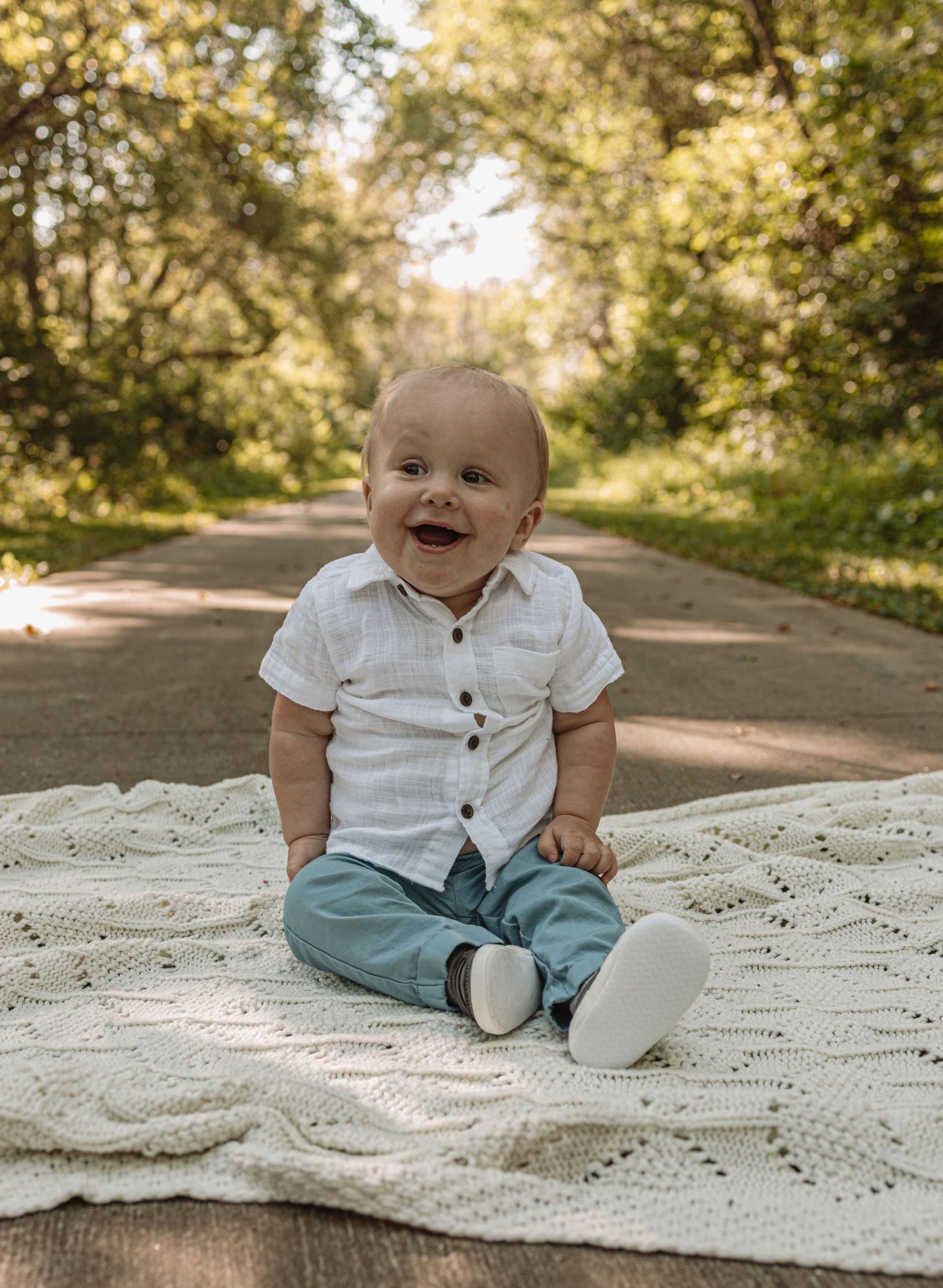 Baby sits on white lace blanket outdoors wearing white button-up shirt and teal pants in autumn setting.