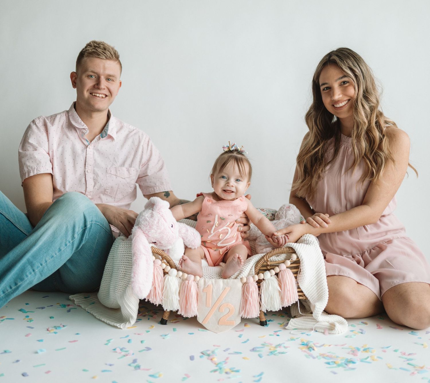 A family in coordinating pink outfits sits together on a confetti-covered floor with their baby in a decorated basket.