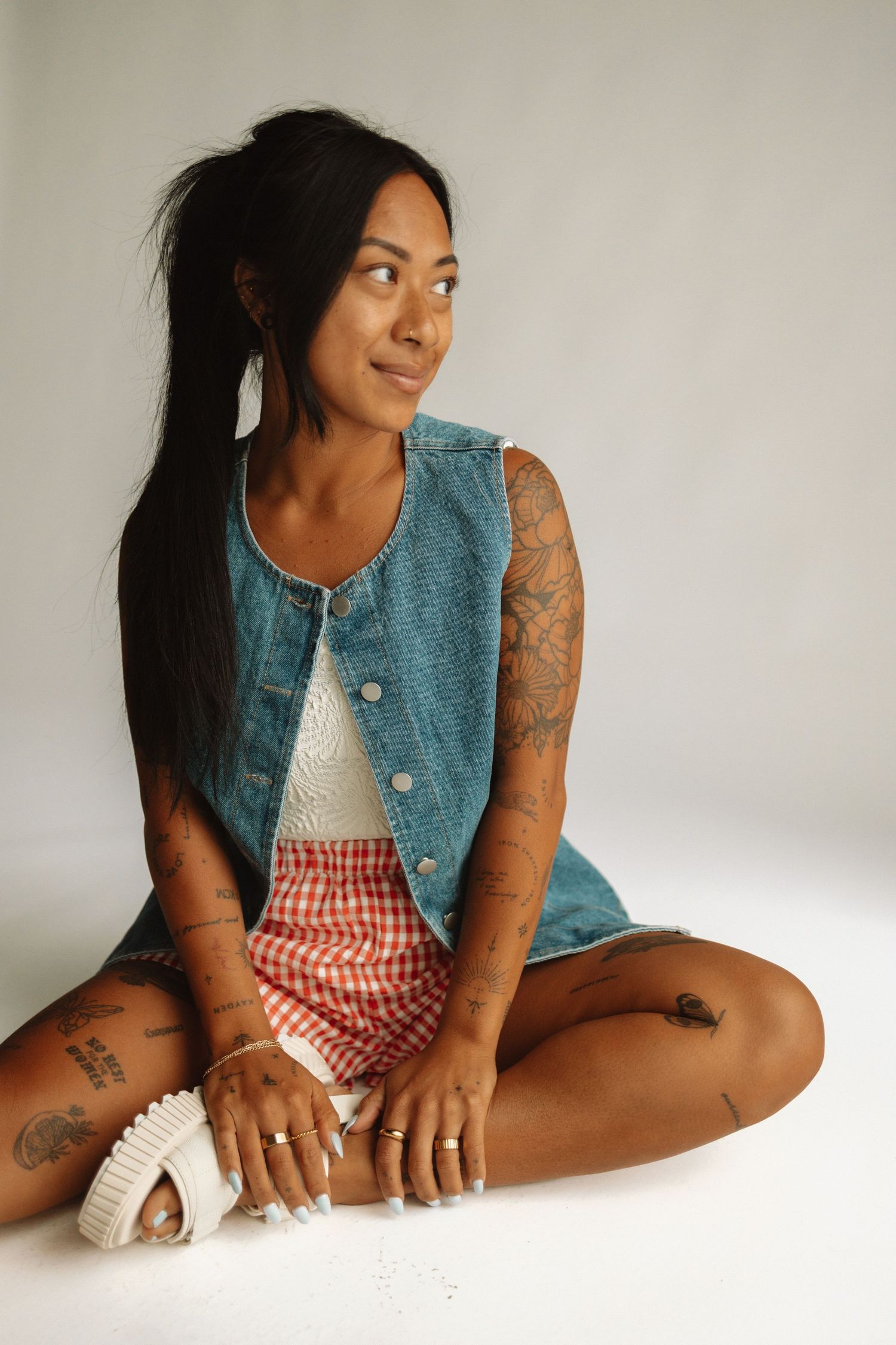 Person in denim vest and red gingham shorts sits casually on white backdrop with tattoos and white sneakers.