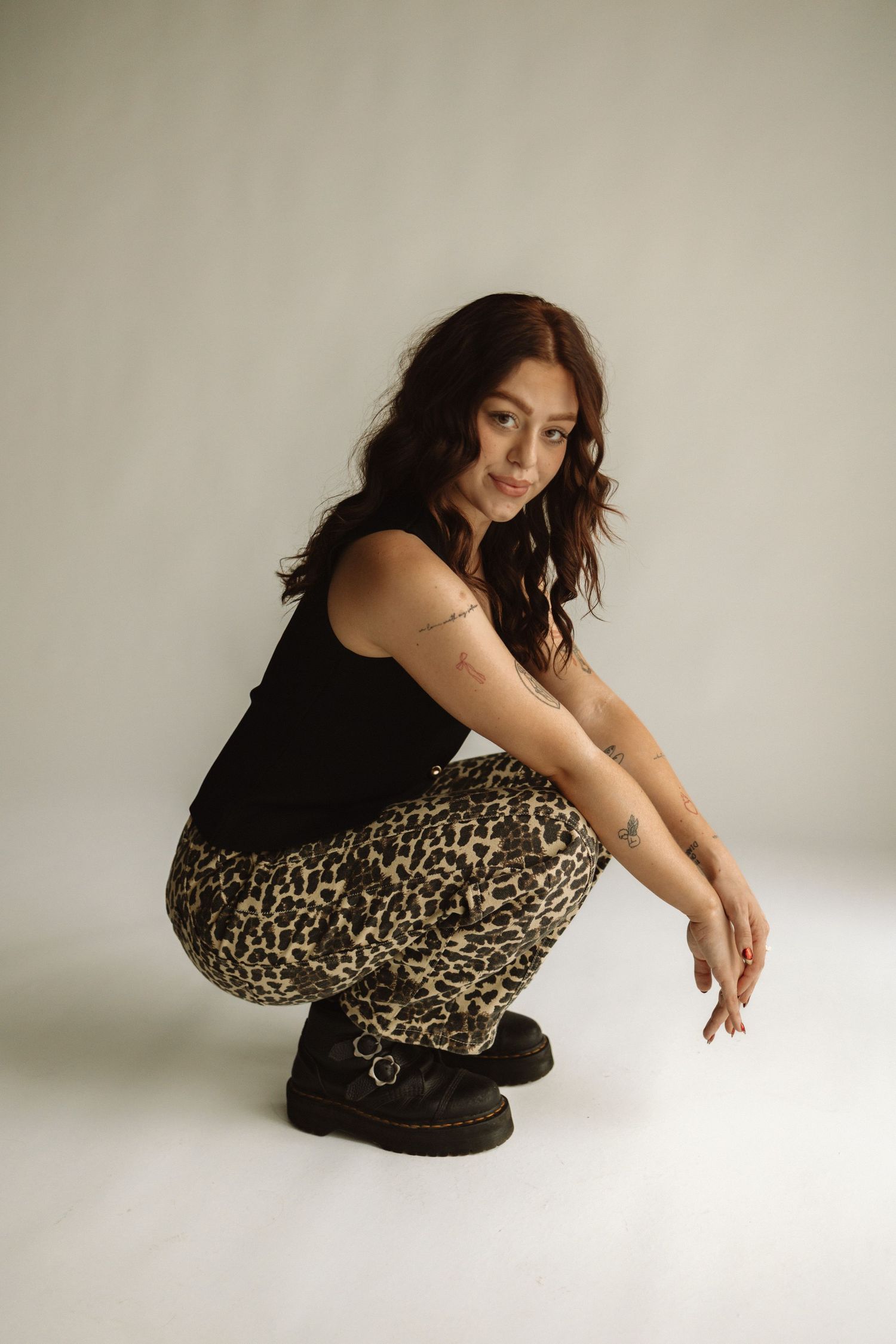 Studio portrait of someone crouching in a black tank top, leopard print pants and platform boots against white background.
