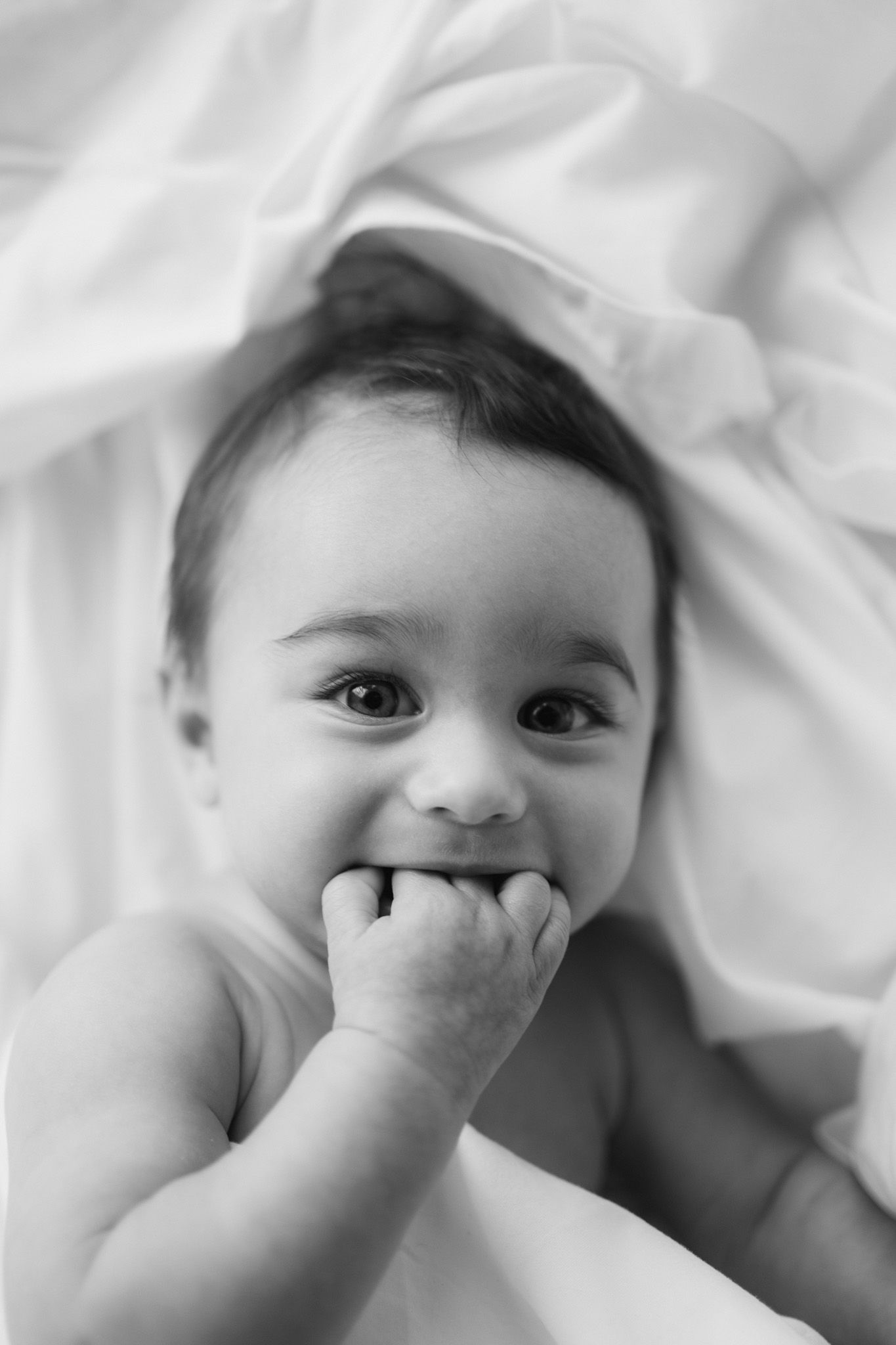 A baby with bright eyes chews on their fingers while lying on white sheets.