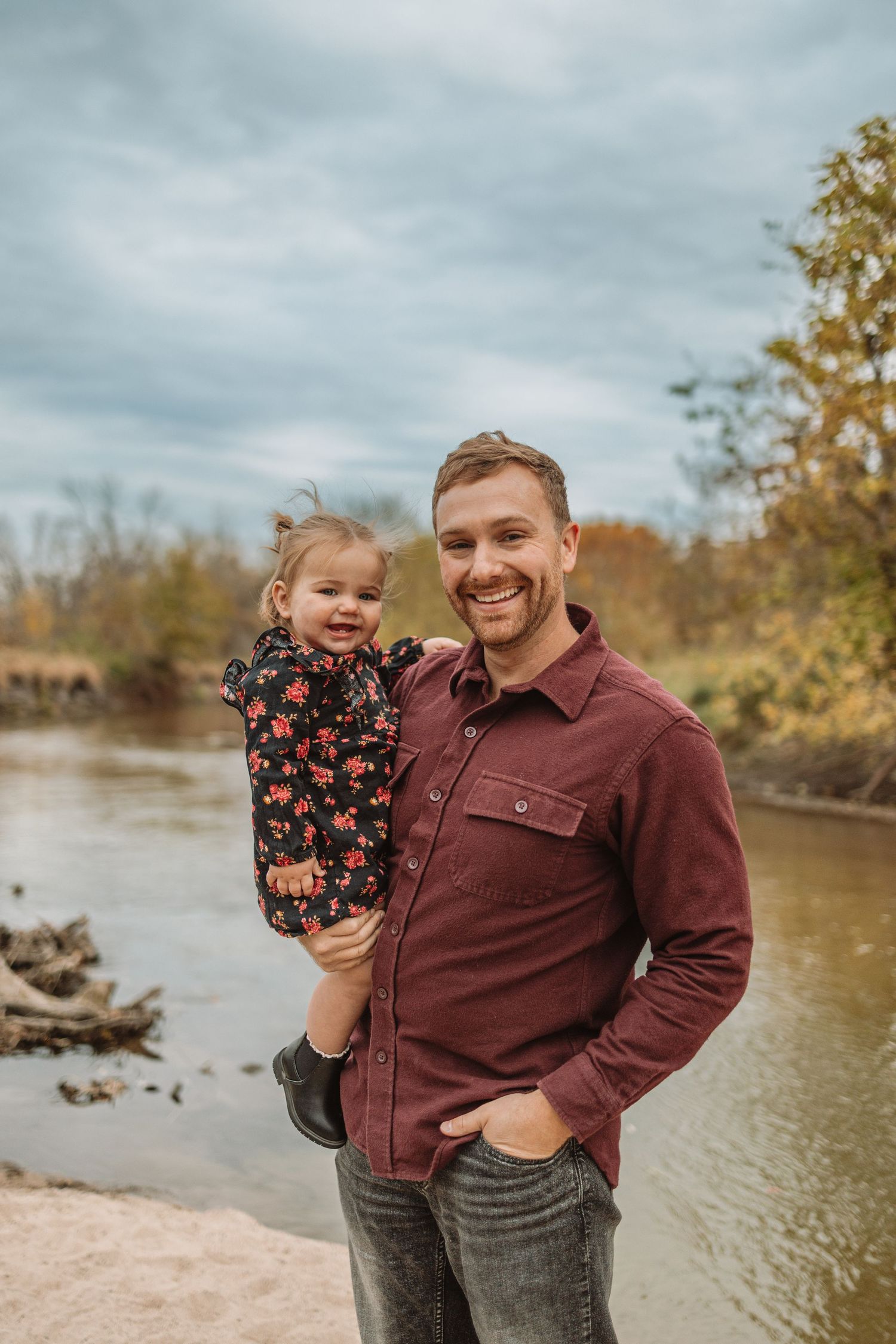 A parent and child share a joyful moment together by a river on an autumn day with cloudy skies.