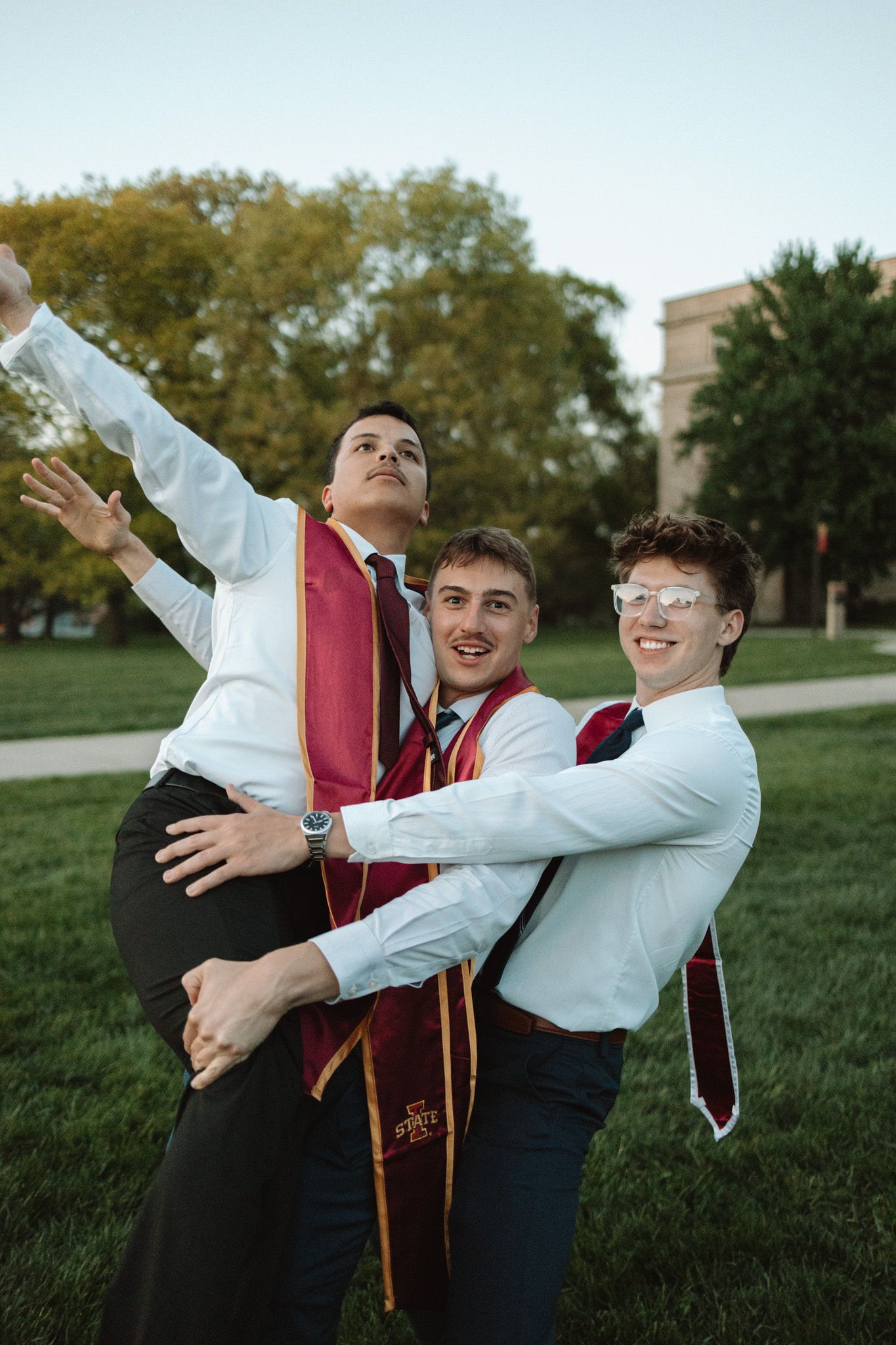 Three graduates in formal attire pose playfully on a college campus lawn with red stoles and white shirts.