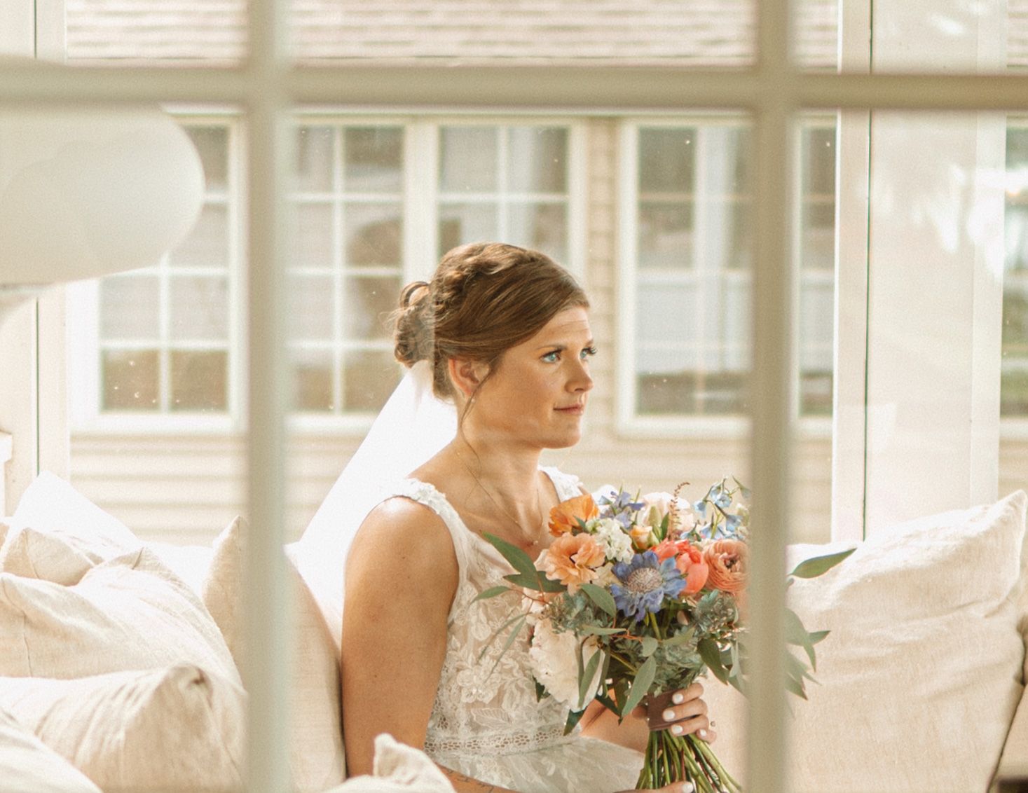 A bride in a stunning white lace wedding dress carries a vibrant peach and orange bouquet while walking in natural light.