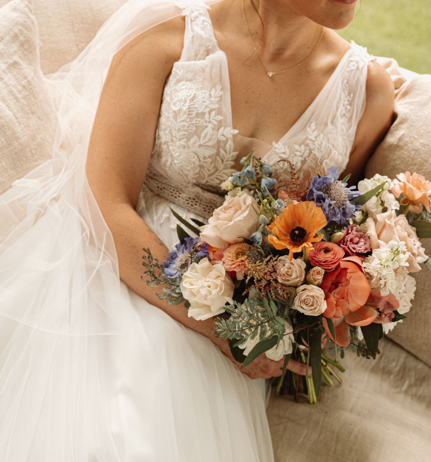 A wedding bouquet with peach roses, white flowers, and purple accents rests on a white lace dress with floral details.