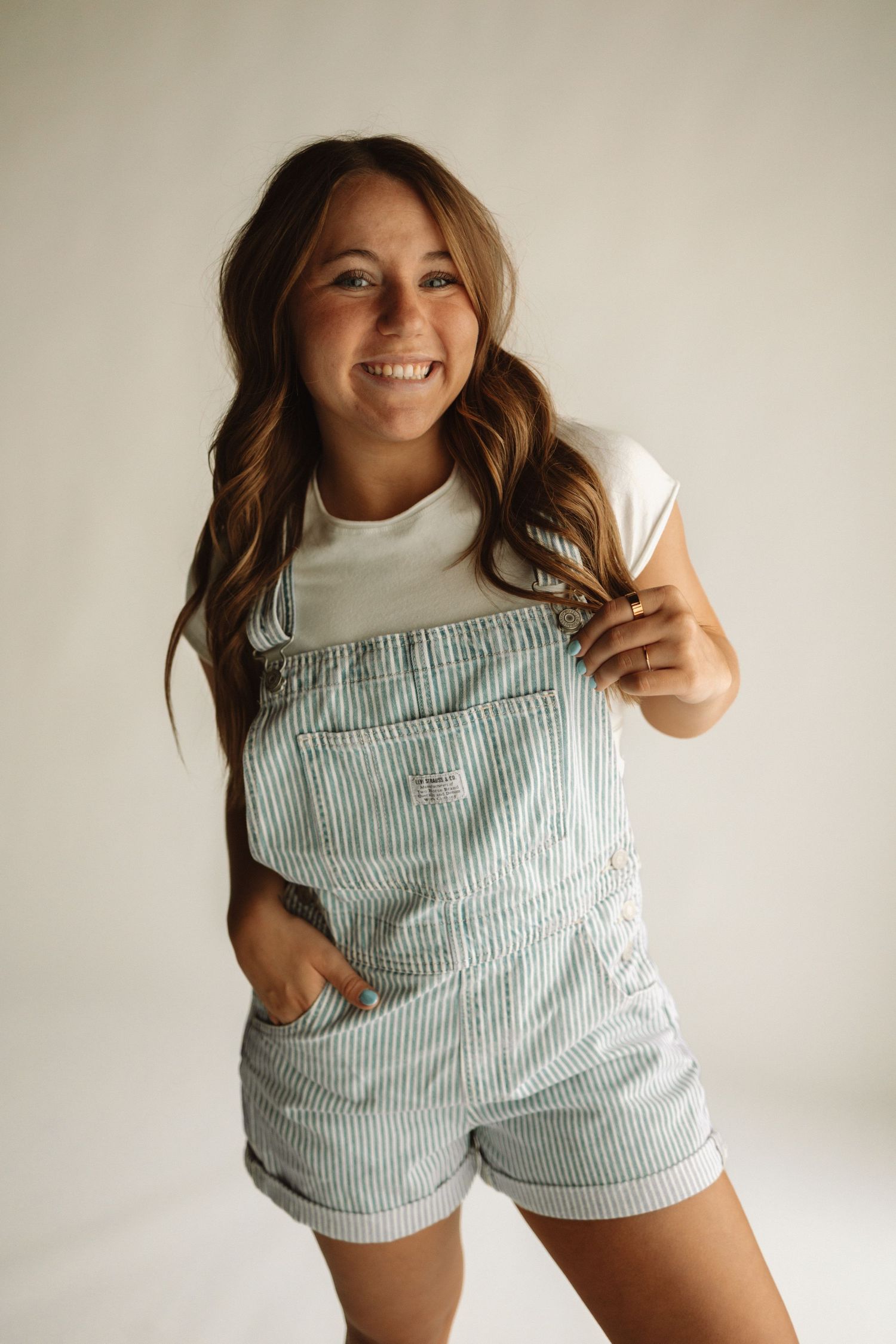 Young person wearing white turtleneck and light blue striped overalls shorts poses playfully in studio setting.