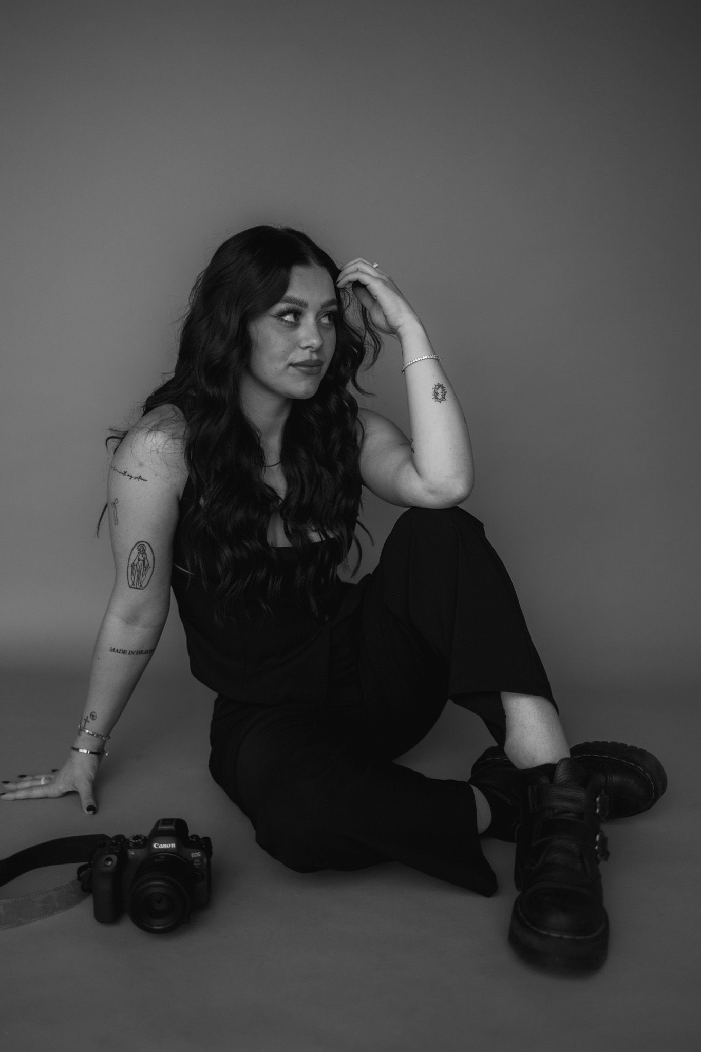 A black and white portrait of a photographer sitting on the floor with a camera and wearing dark clothing.
