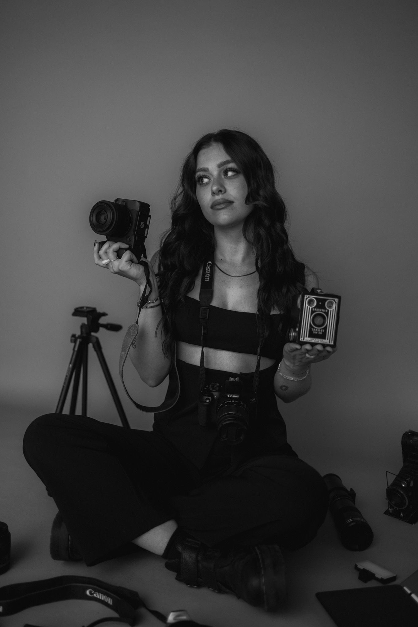 Monochrome photo of a photographer holding vintage cameras while sitting on the floor surrounded by equipment.