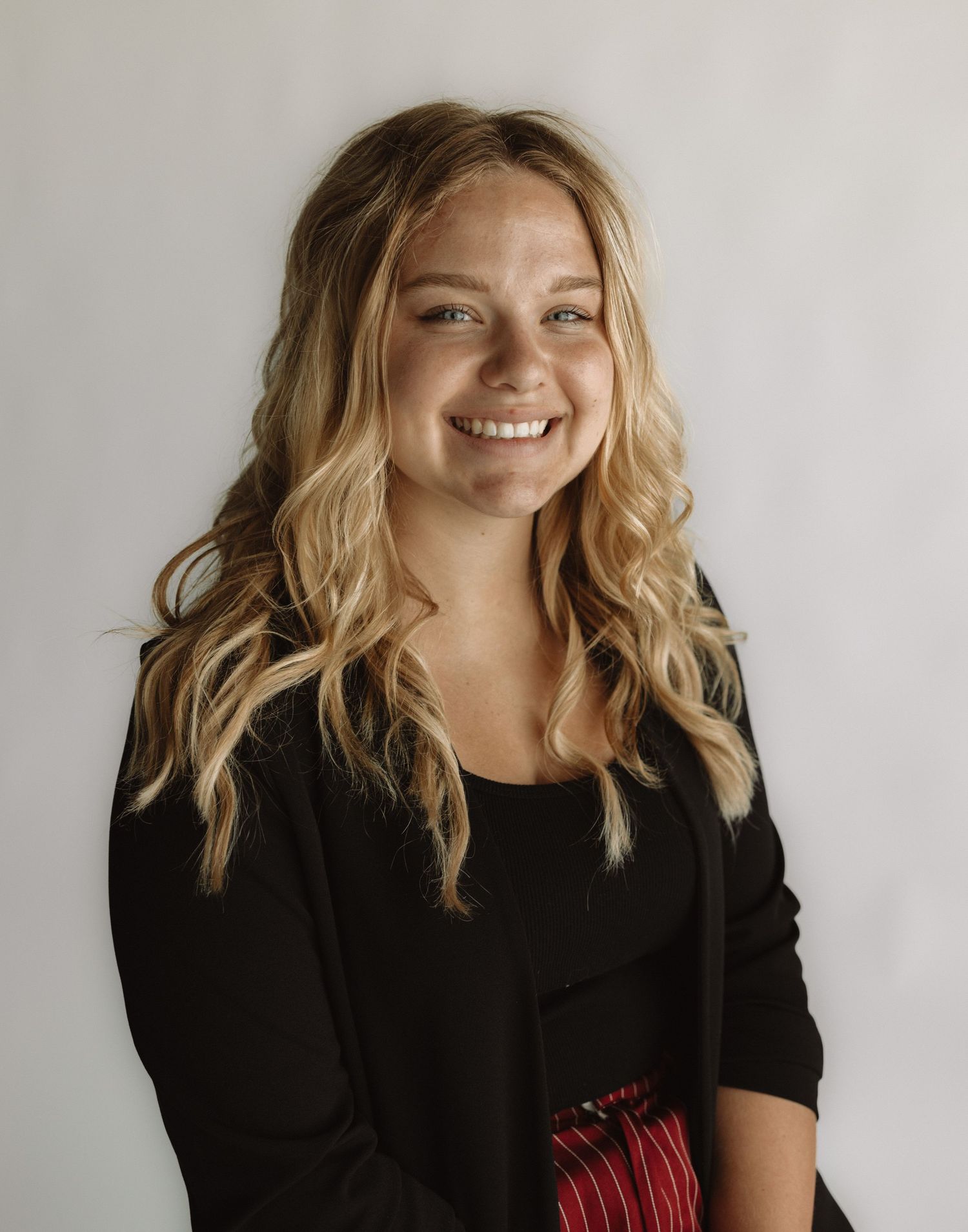 A person with wavy blonde hair wearing a black top smiles warmly against a light background.