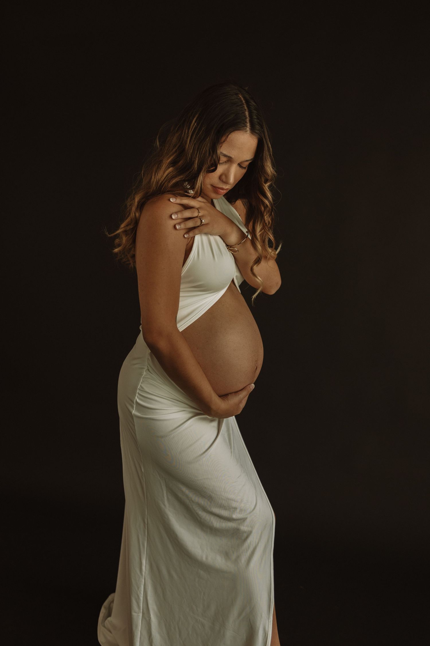 Pregnancy maternity photoshoot featuring woman in elegant white dress against dark background with artistic side angles.