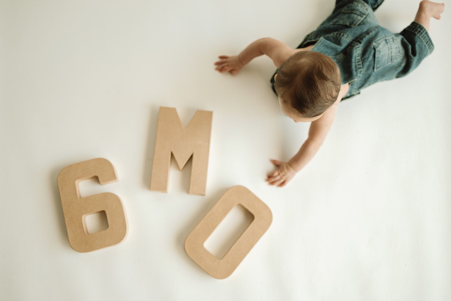 A young child lying on a white background with wooden numbers and letters spelling out 6 months.