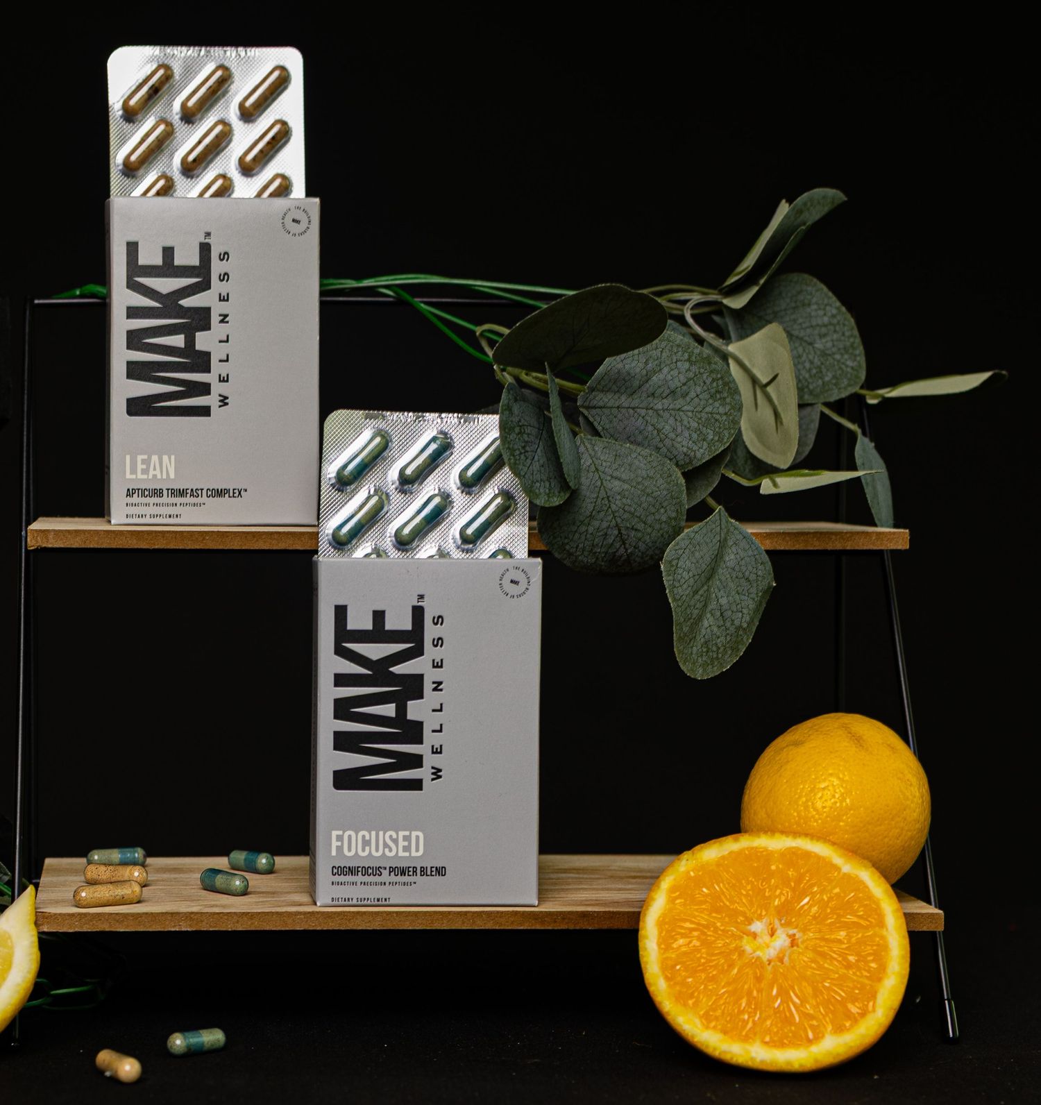 Two MAKE brand product boxes displayed on wooden shelves with fresh oranges and eucalyptus leaves against dark background.