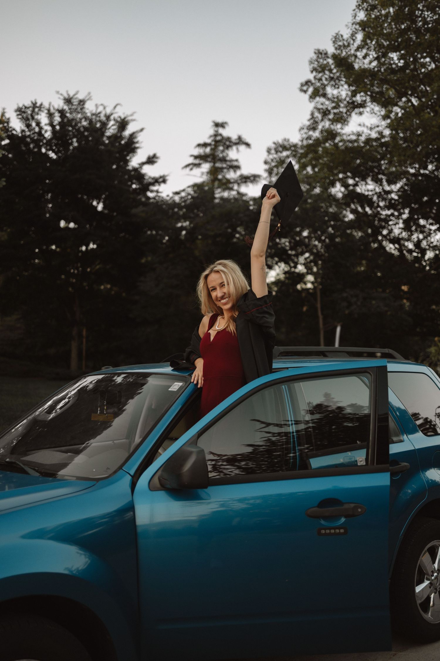 A person in a red top poses joyfully through the sunroof of a bright blue car at sunset.