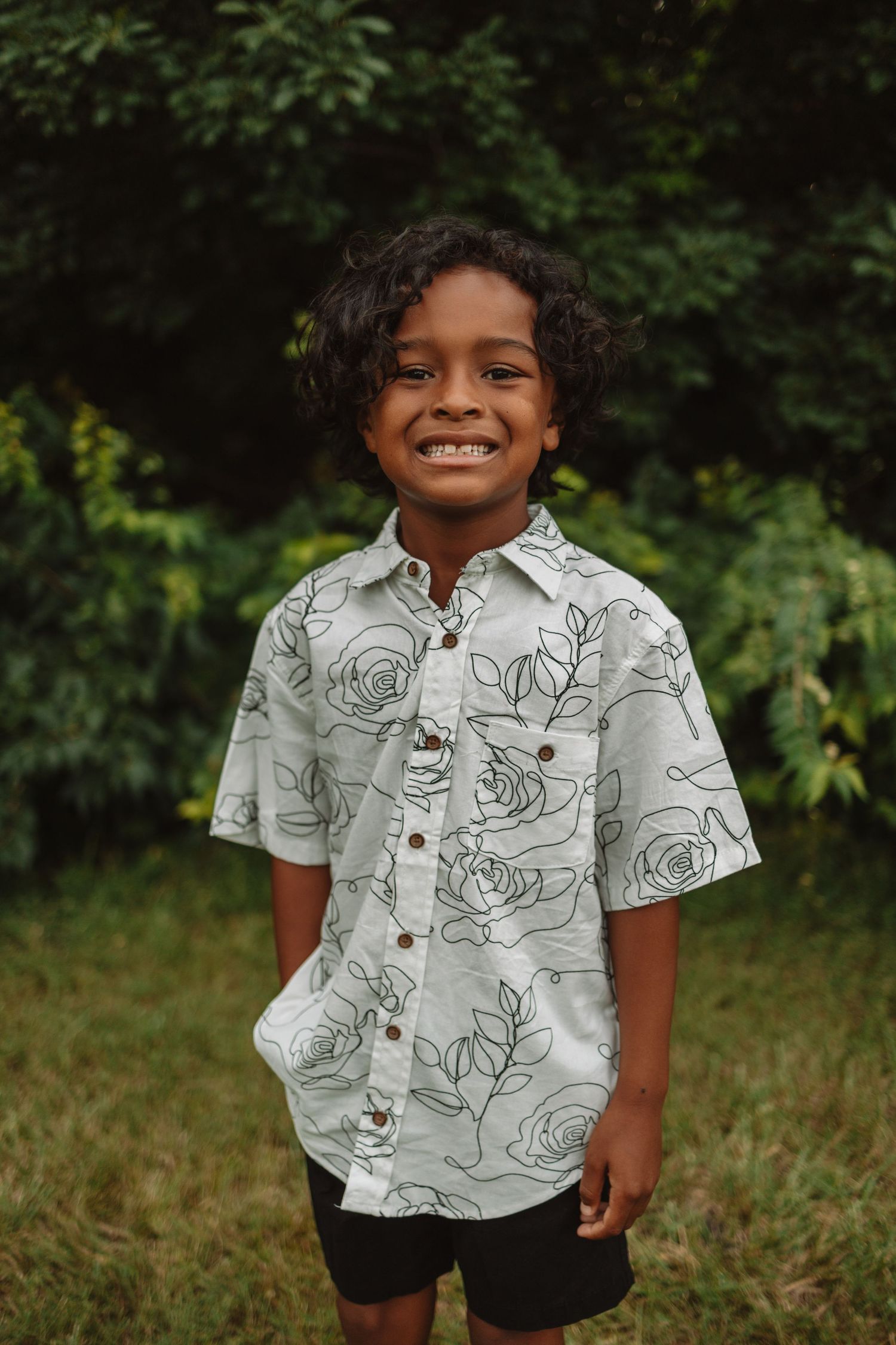 Young child wearing a white button-up shirt with black floral print design poses outdoors in grass and trees.