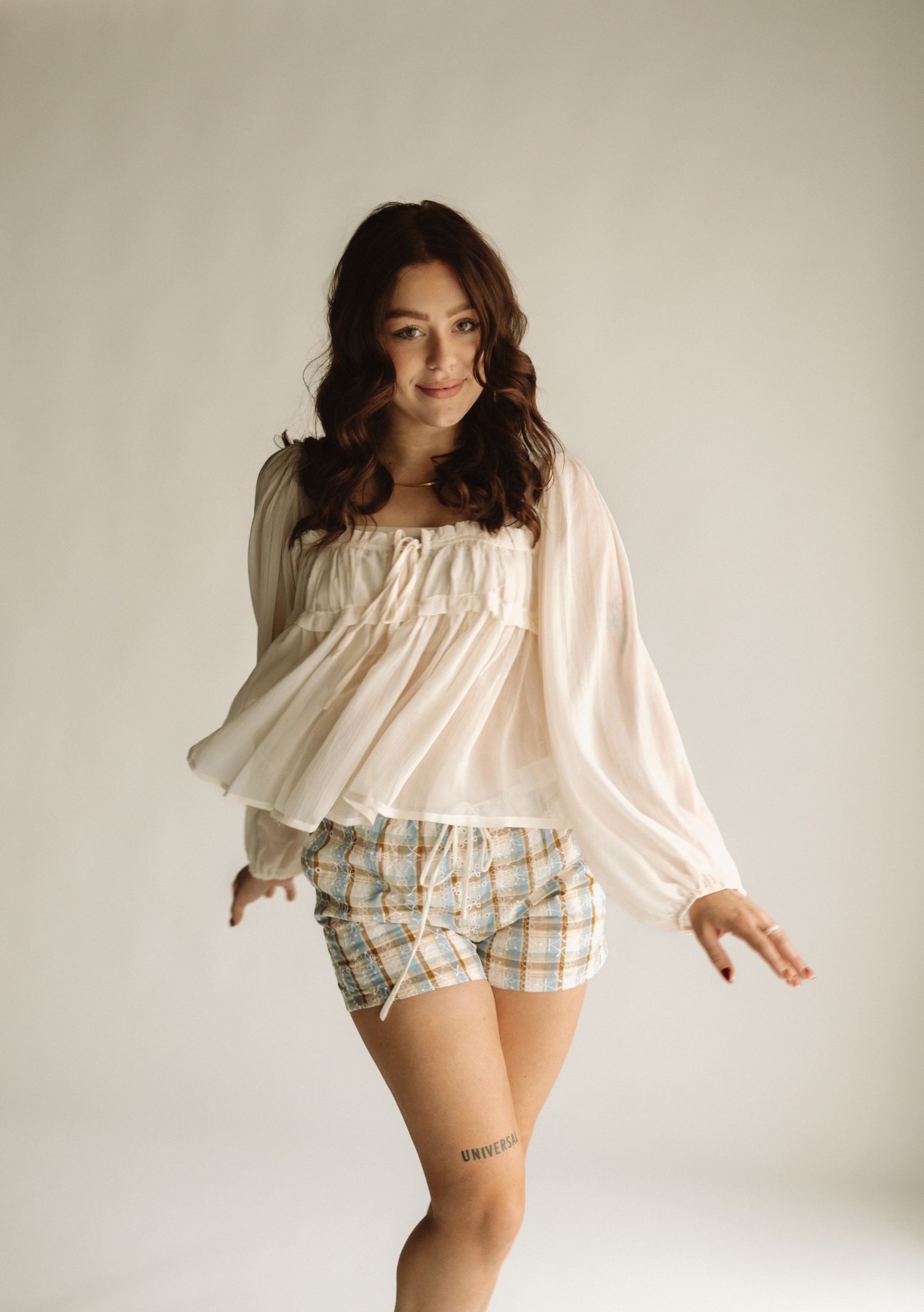 Figure in flowy white top and plaid shorts poses playfully against neutral backdrop in lifestyle photoshoot series.