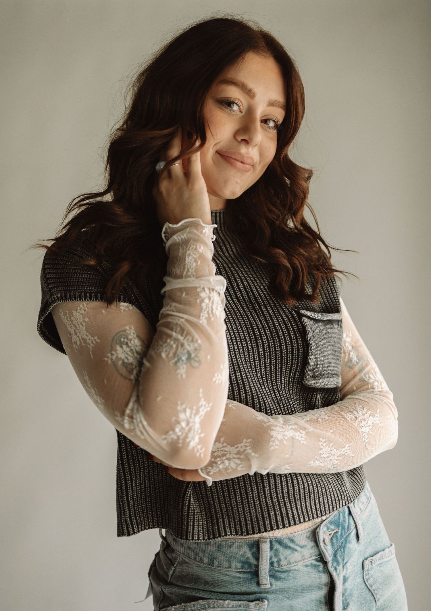 A person wearing a gray knit vest over a white lace long sleeve top poses against a neutral background.