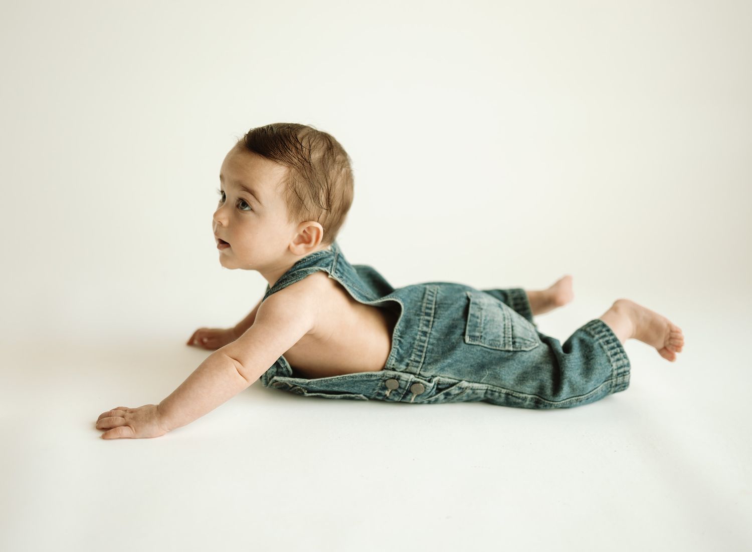 Baby in teal overalls lying on stomach against white background.
