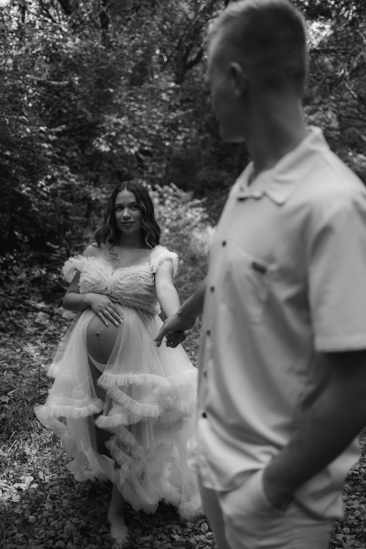 Black and white outdoor maternity photoshoot showing couple in ethereal white dress and casual attire amid foliage.
